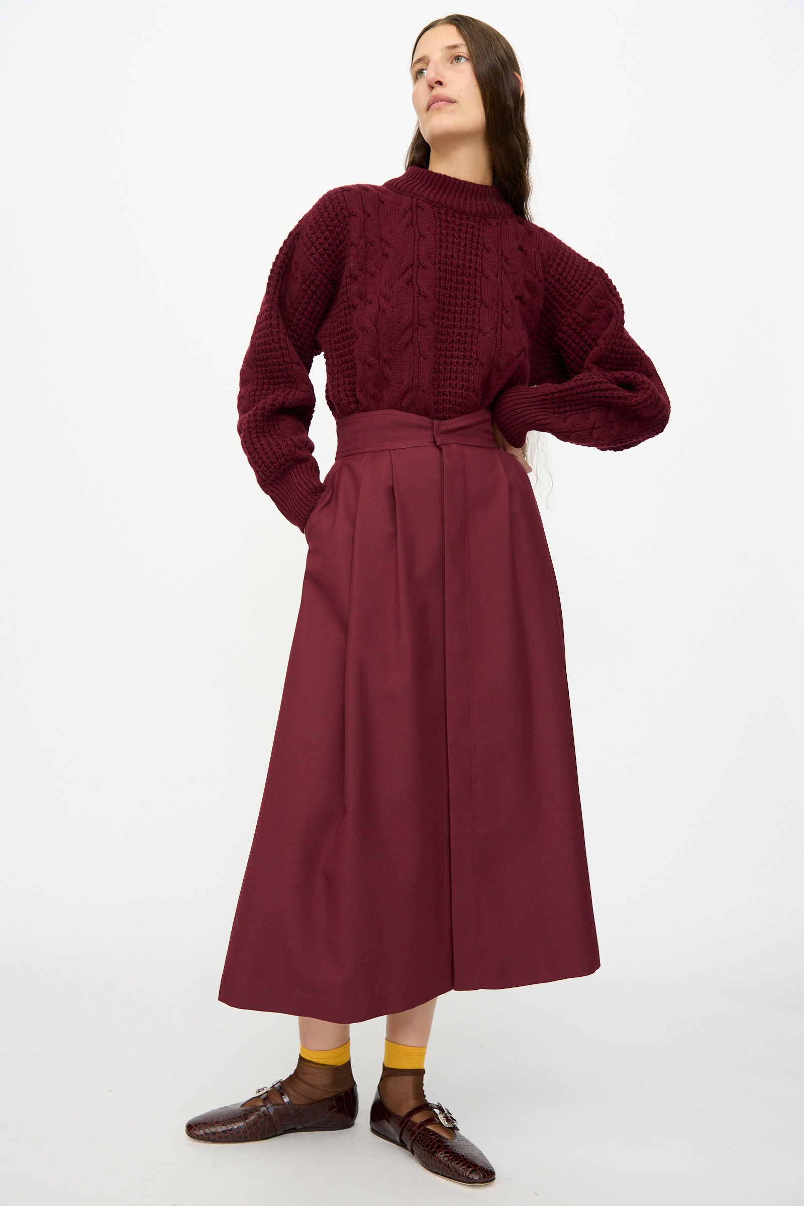 A woman stands against a white background wearing a textured burgundy sweater, yellow socks, brown shoes, and the Les Prairies De Paris Twill Georgie Skirt in Bordeaux.