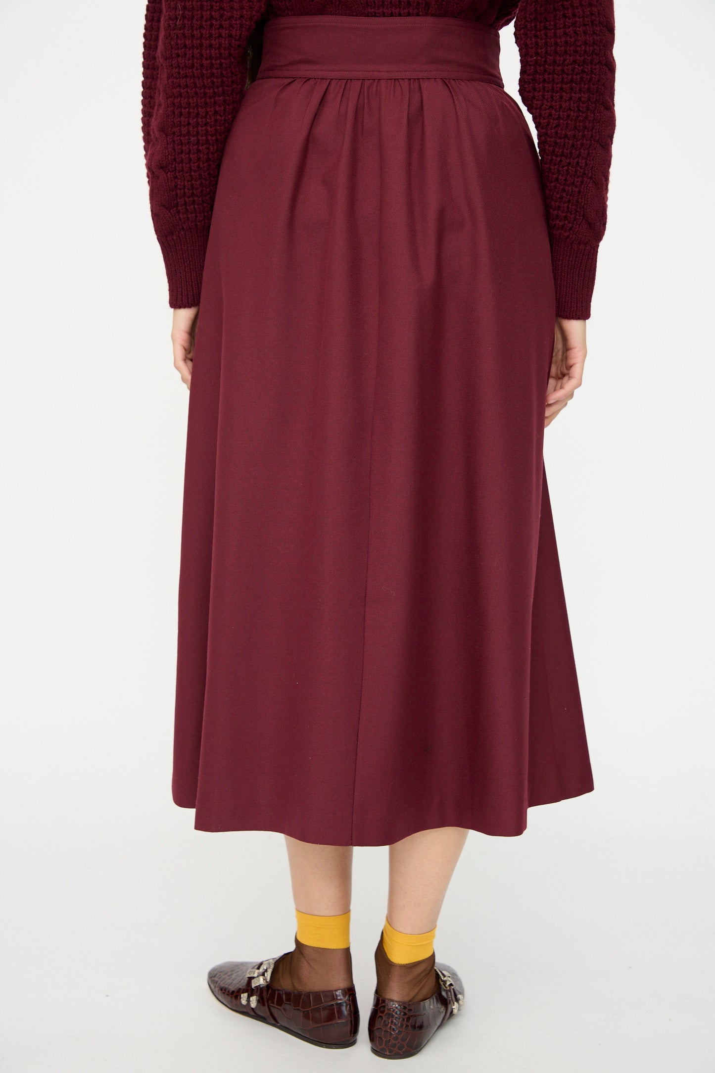 A person wears the Les Prairies De Paris Twill Georgie Skirt in Bordeaux with a matching sweater, yellow socks, and brown shoes, seen from behind against a plain background.