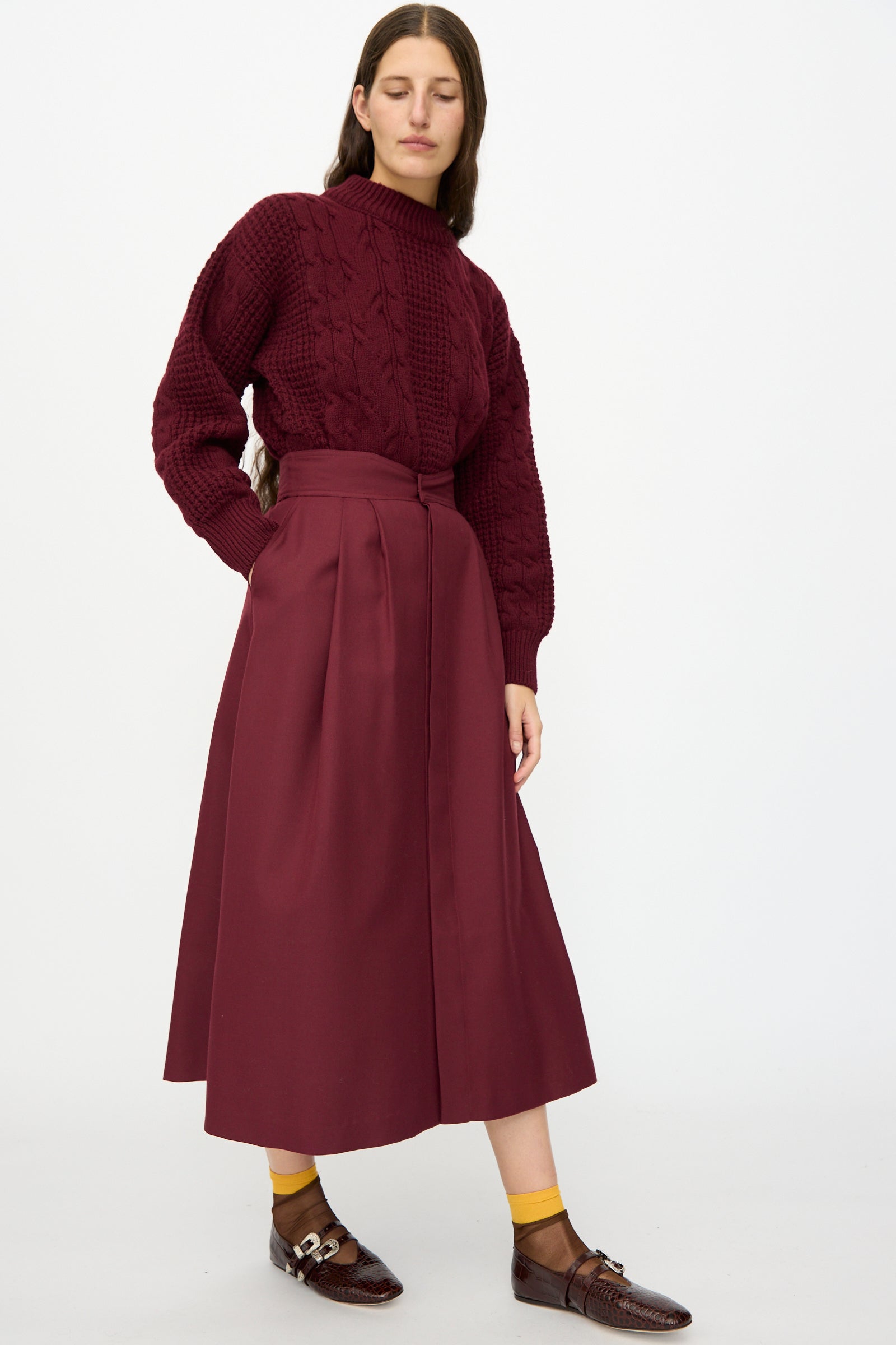 A woman wears the Les Prairies De Paris Twill Georgie Skirt in Bordeaux with a textured burgundy sweater, mustard socks, and brown loafers, standing against a plain white background.