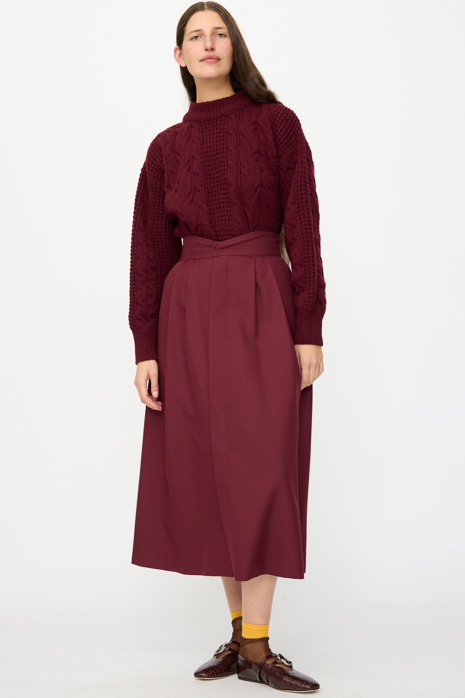 A woman stands against a white background wearing the Les Prairies De Paris Twill Georgie Skirt in Bordeaux, paired with a maroon cable-knit sweater, brown shoes, and yellow socks.