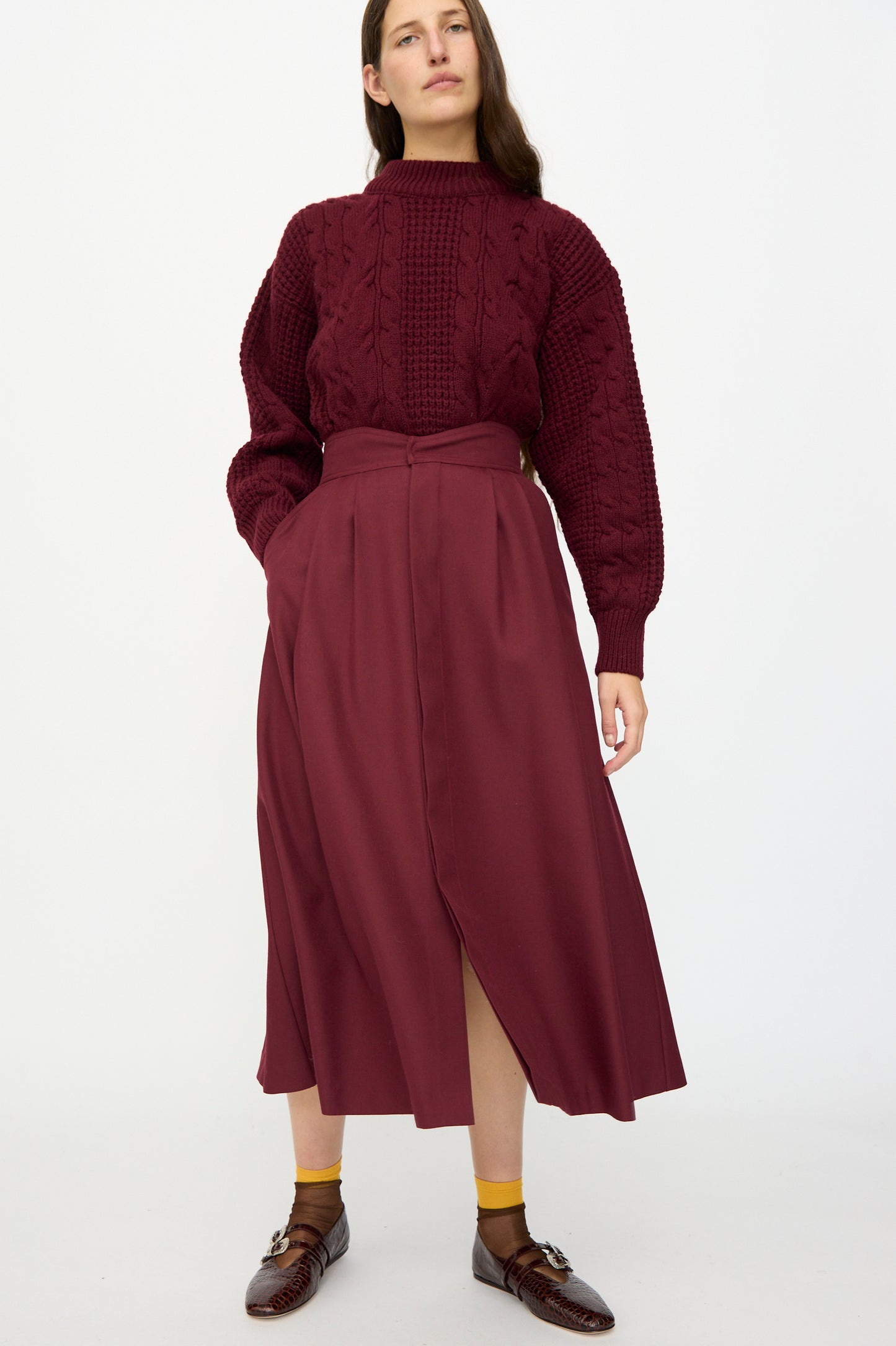 A person stands against a white background wearing the Les Prairies De Paris Twill Georgie Skirt in Bordeaux, styled with a textured burgundy sweater, yellow socks, and brown dress shoes. Made in France.