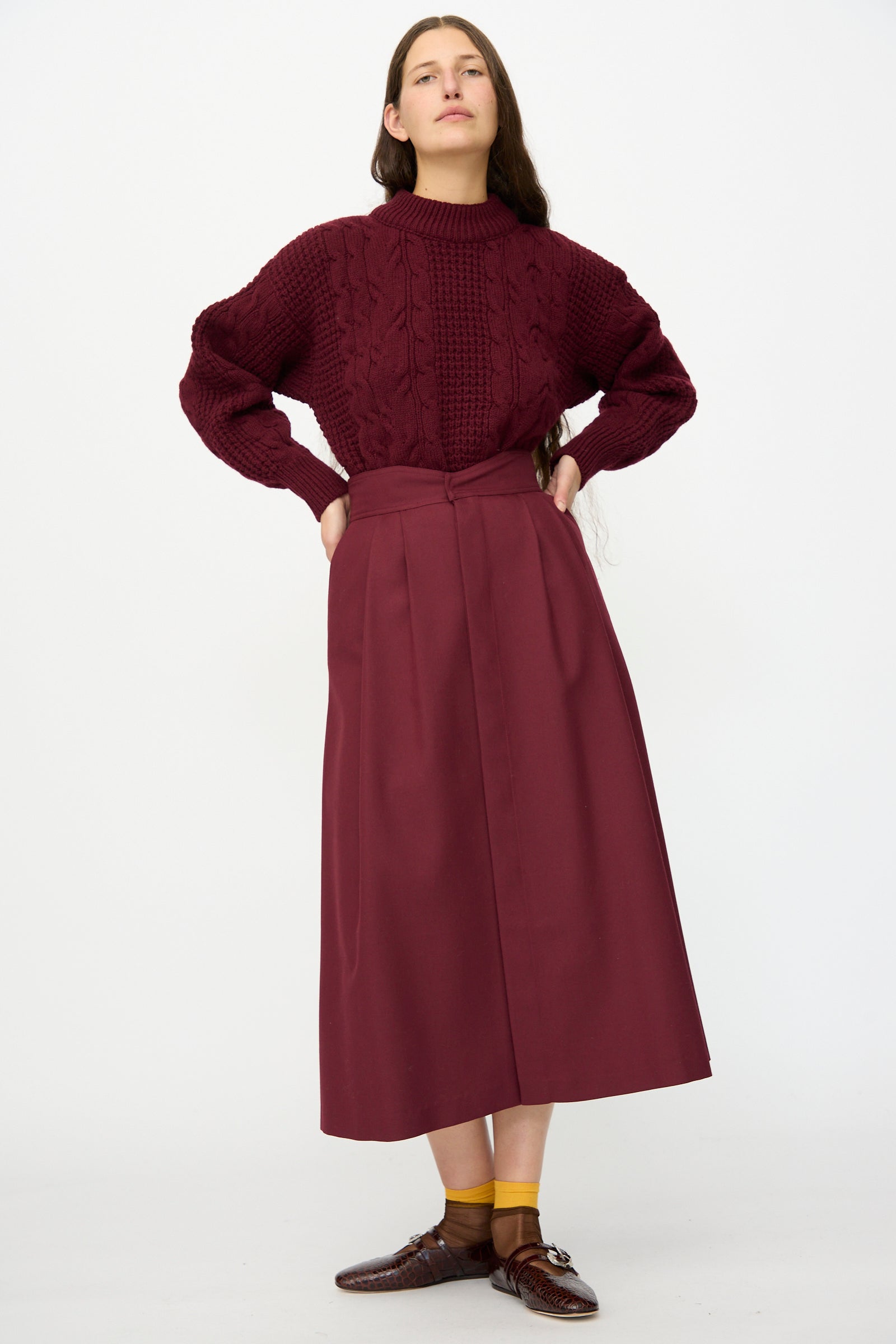 A woman stands against a white background wearing a maroon cable-knit sweater, the Les Prairies De Paris Twill Georgie Skirt in Bordeaux, brown shoes, and yellow socks.