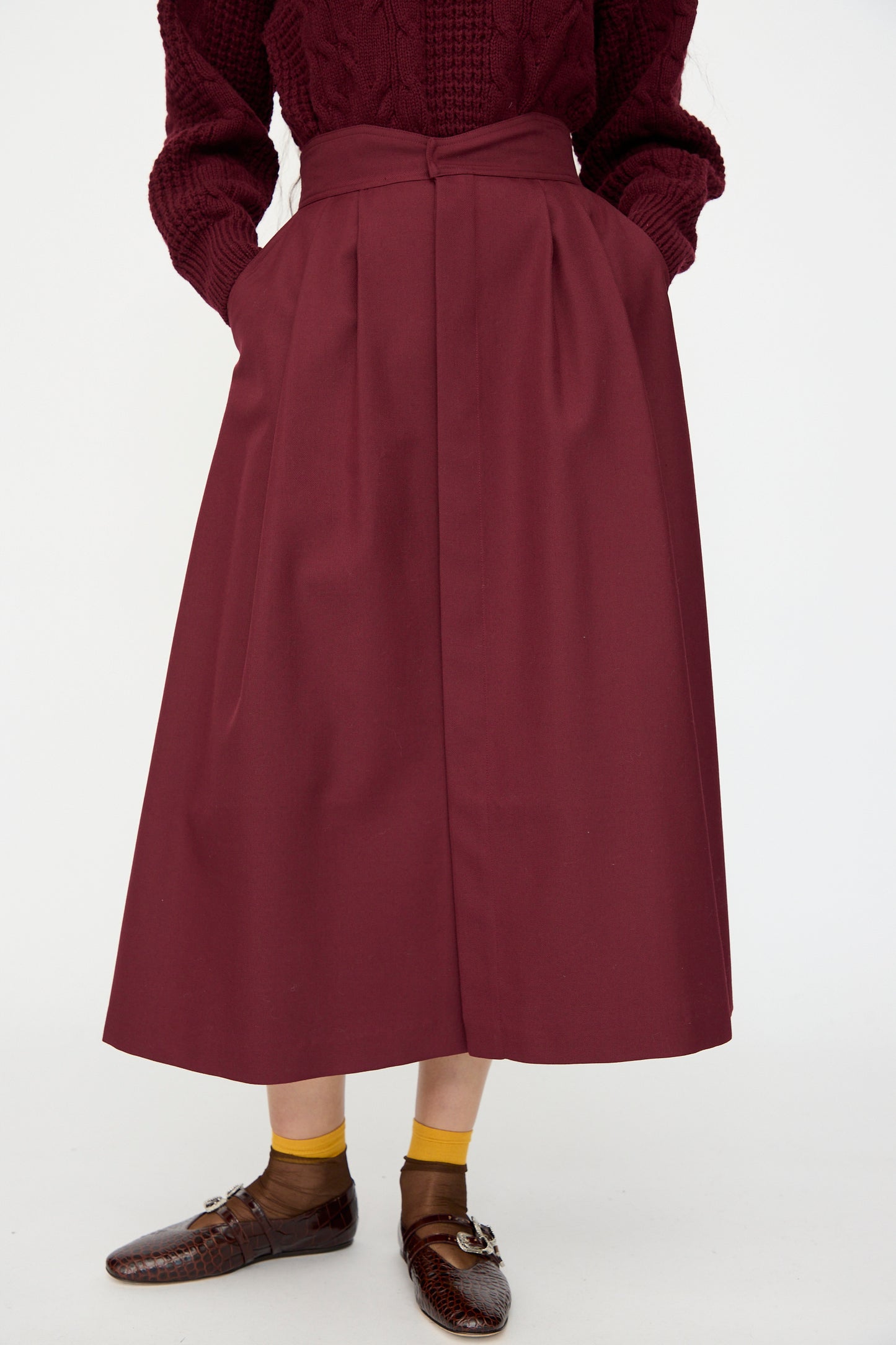 A person wearing the Les Prairies De Paris Twill Georgie Skirt in Bordeaux with a matching sweater, yellow socks, and brown shoes stands against a plain white background.