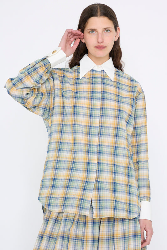 A person wearing a plaid outfit with a white collar and cuffs layers it over the Chemise Oxford in Paper Miel by Les Prairies De Paris, standing serenely against a plain white background.