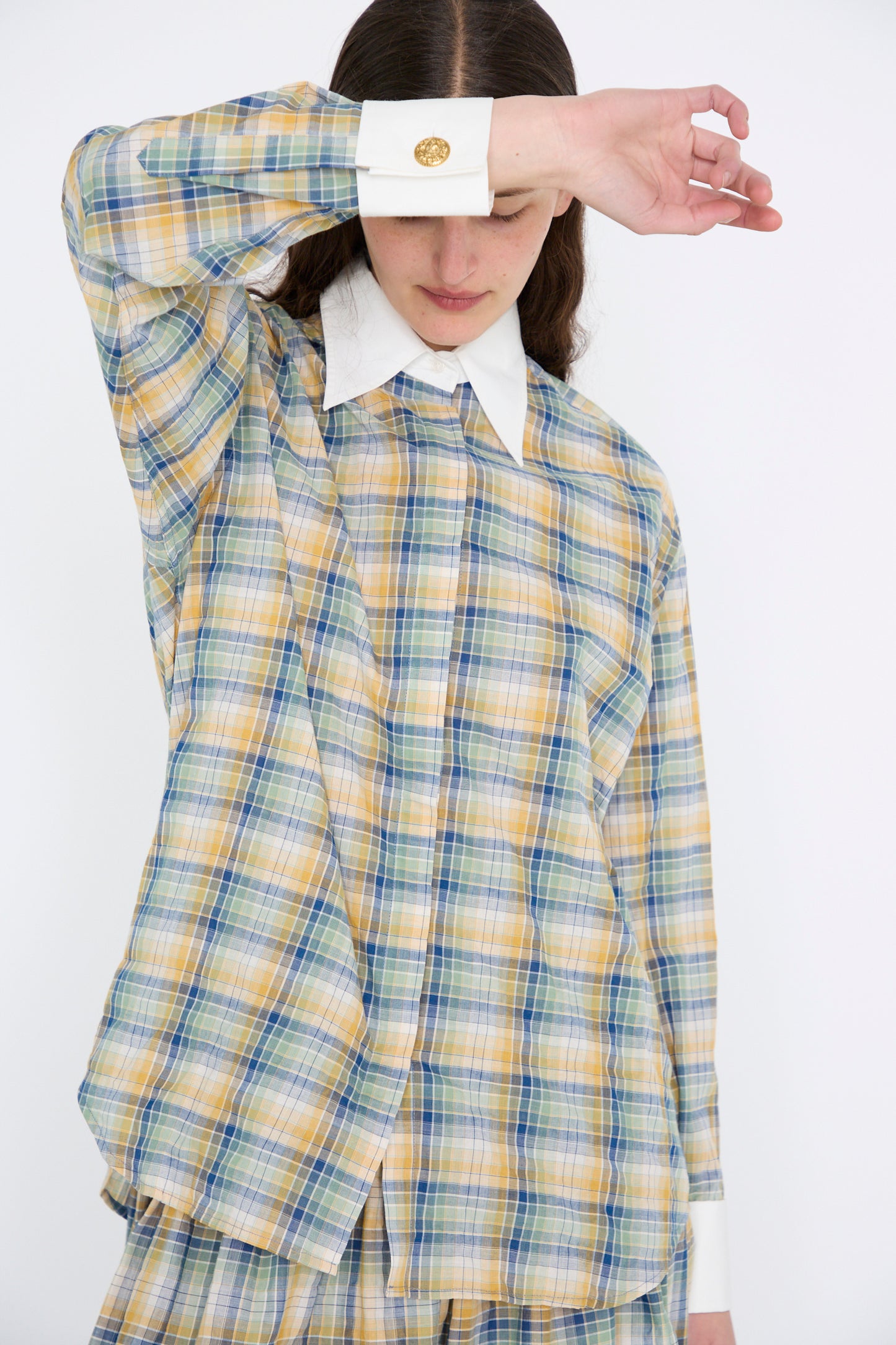 A person wearing the Chemise Oxford in Paper Miel by Les Prairies De Paris, paired with matching pants and gold cufflinks, shields their face with one arm against a plain backdrop.