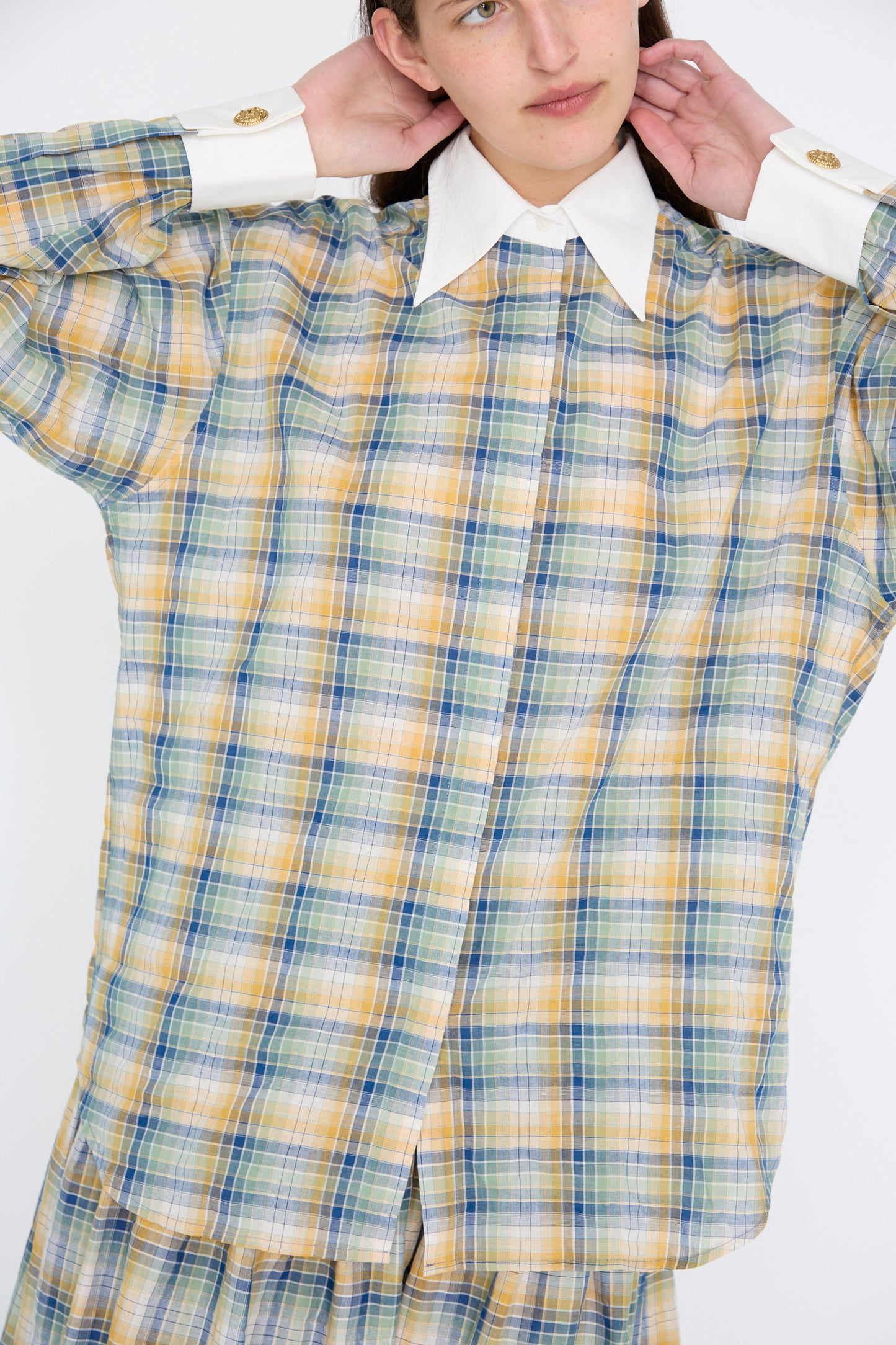 A person is wearing the loose-fitting Chemise Oxford in Paper Miel by Les Prairies De Paris, featuring a multicolored plaid design with a white collar and cuffs, made from lightweight striped cotton, posing with hands behind their neck.