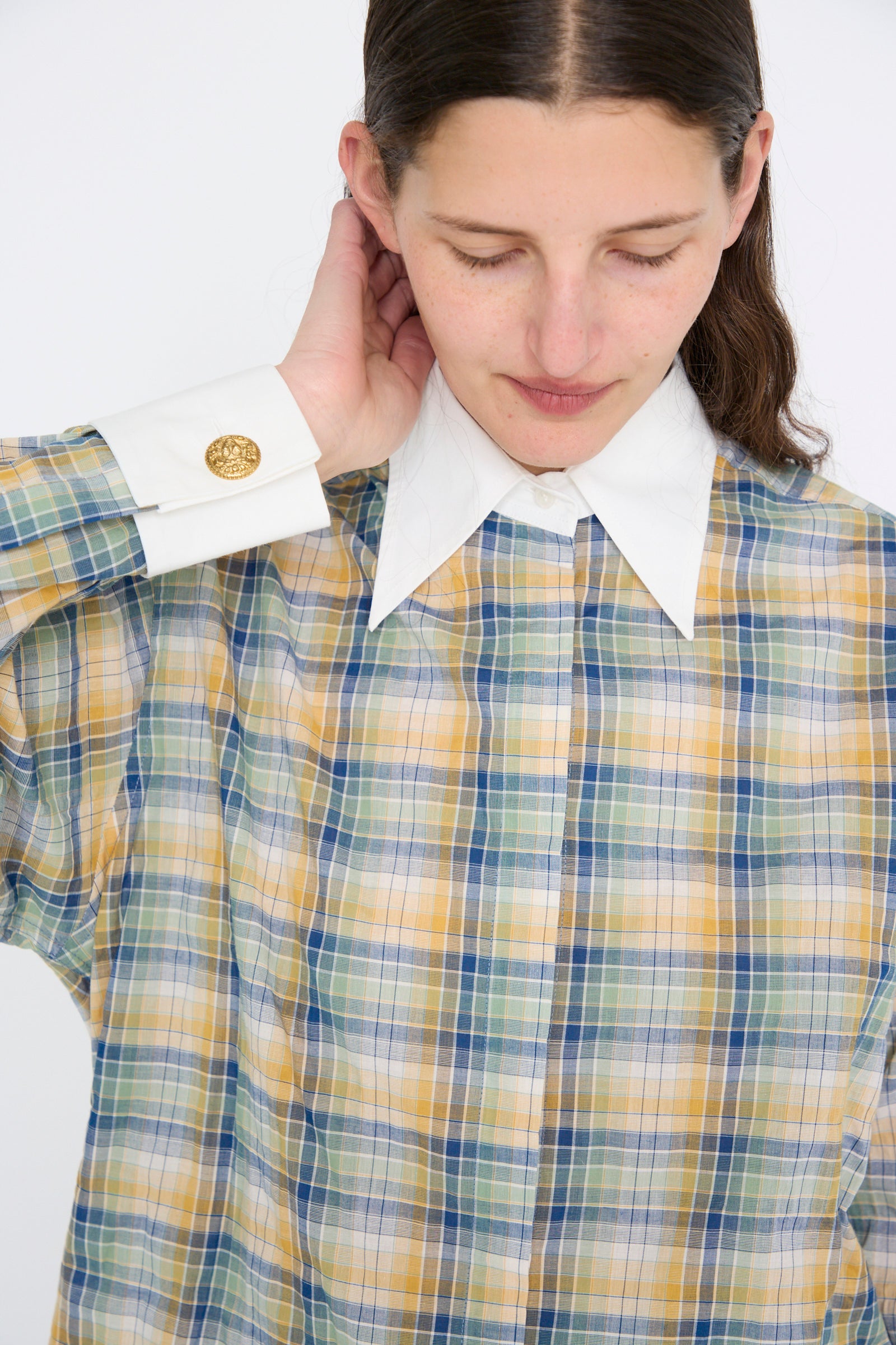 A person in the Chemise Oxford in Paper Miel by Les Prairies De Paris, featuring a plaid pattern with a large white collar and gold-buttoned cuffs, looks down and touches their neck. The lightweight striped cotton fabric adds a breezy touch to the classic design.