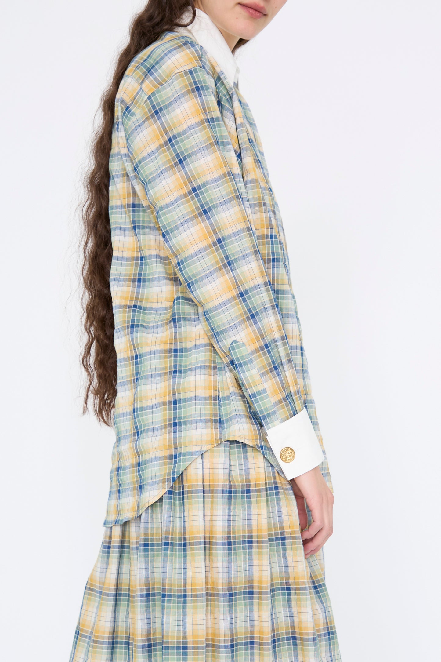 A person with long hair is wearing Les Prairies De Paris' Chemise Oxford in Paper Miel, a plaid shirt in yellow, green, and blue, featuring a white collar and cuffs, over a matching skirt.