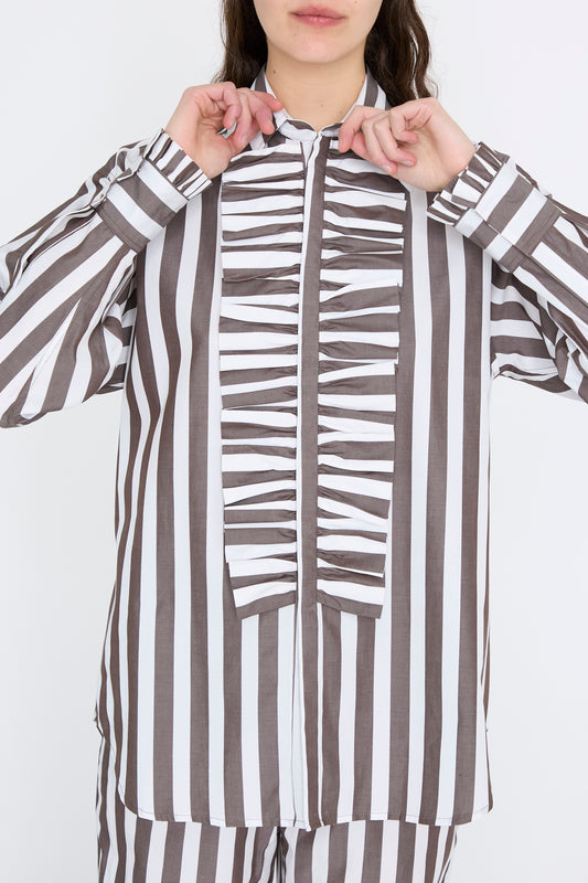 A model wears the "Chemise Serge in Marron" by Les Prairies De Paris, featuring brown and white vertical stripes, against a white backdrop. 