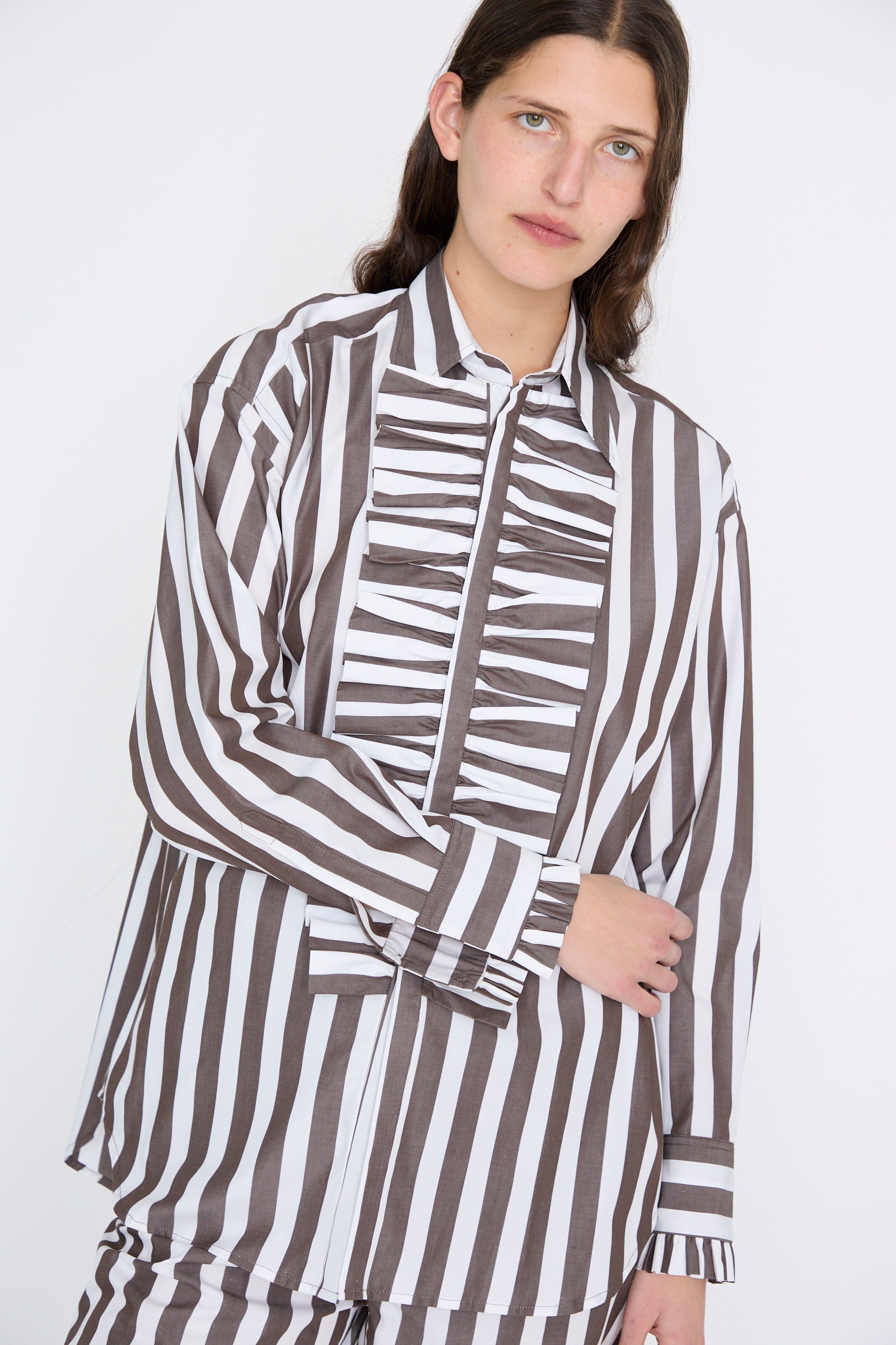 A person wearing the Chemise Serge in Marron, a brown and white vertically striped long sleeve shirt with a pleated front design by Les Prairies De Paris, is looking at the camera.