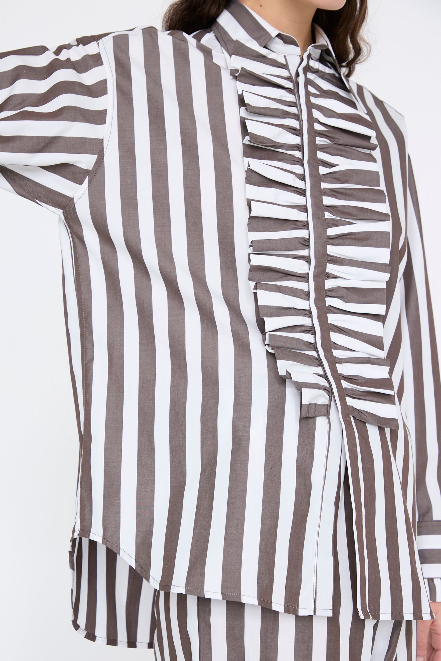 A person in Les Prairies De Paris' Chemise Serge in Marron, featuring vertical brown and white stripes with front ruffles in organic cotton, extends one arm to the side.