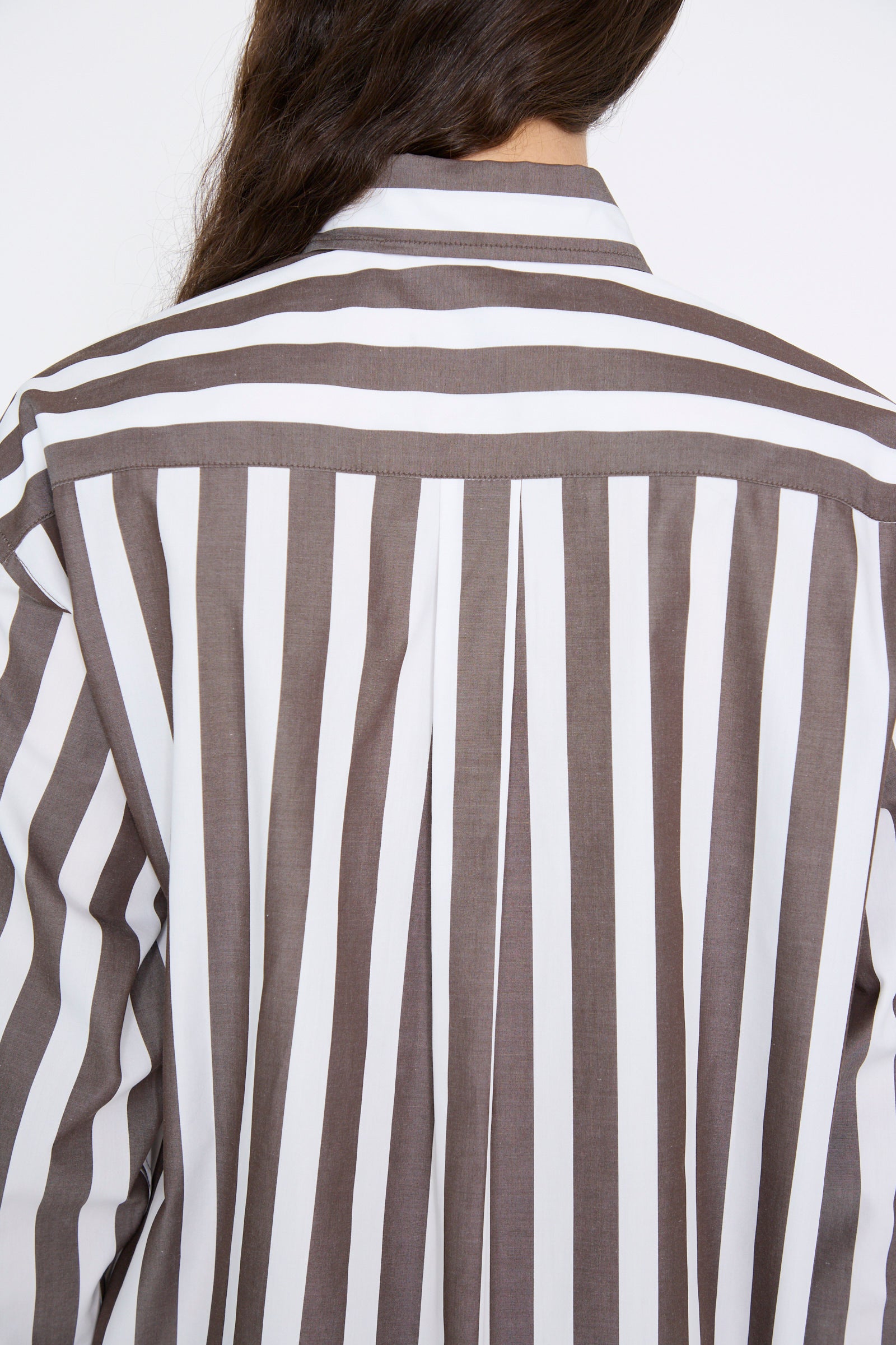 A person with long dark hair is seen from behind wearing the Chemise Serge in Marron by Les Prairies De Paris, a long sleeve shirt featuring vertical brown and white stripes, crafted from organic cotton.