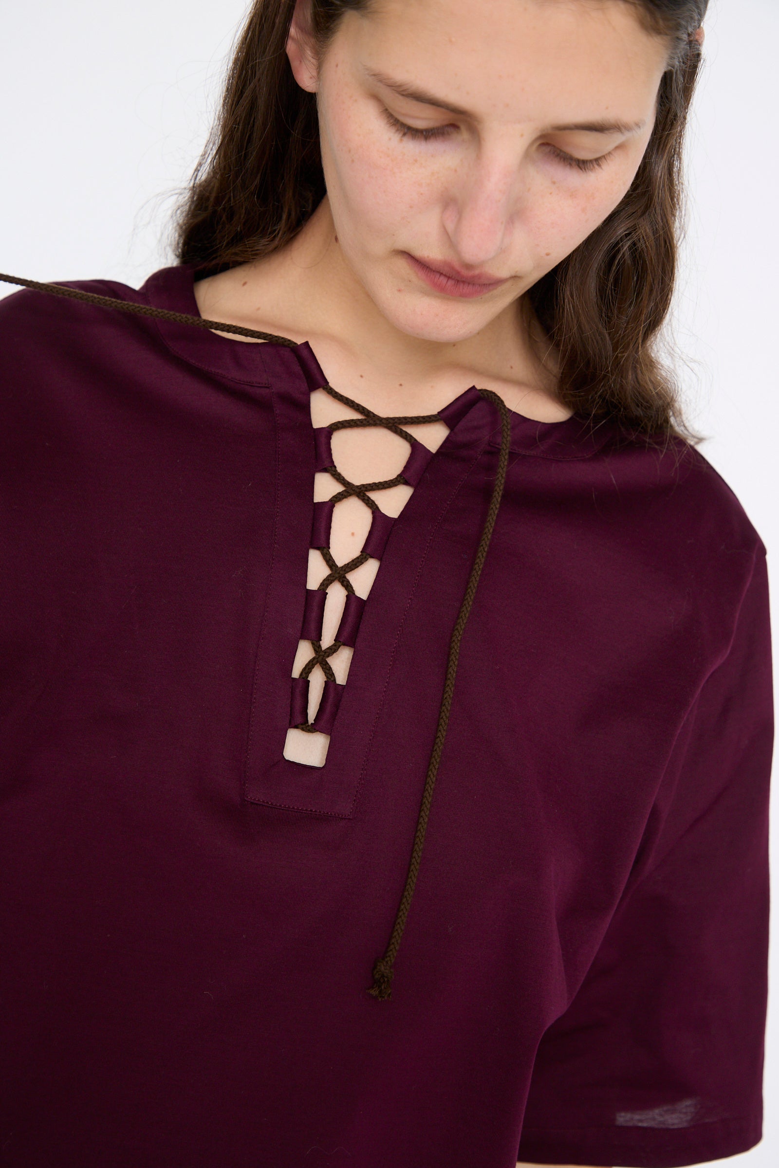 A person wearing the Haut Gaya in Bordeaux by Les Prairies De Paris, a maroon short-sleeve shirt with organic cotton and a crisscross lace-up design near the neckline, looking downward.