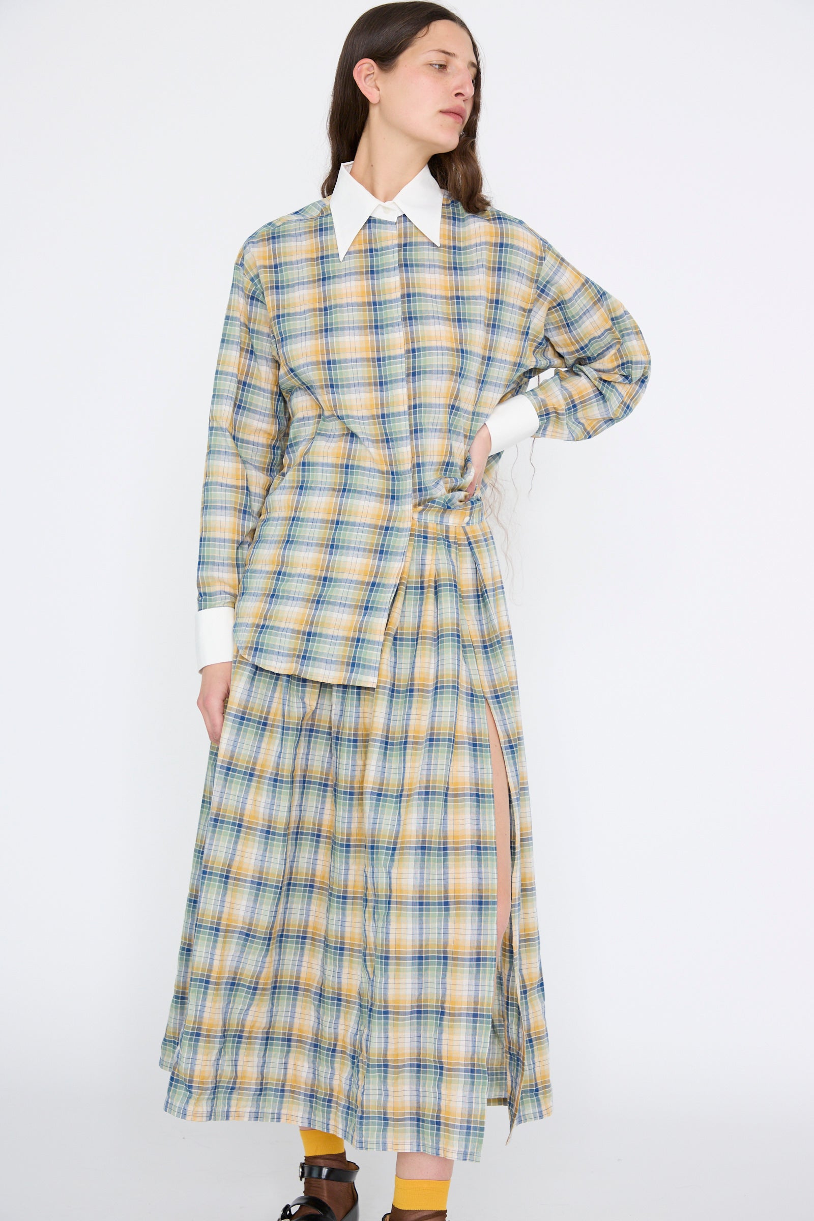 Dressed in Jupe Roberta in Paper Miel by Les Prairies De Paris, featuring a plaid long skirt with yellow, blue, and green checks and a white collar top made from organic cotton, the person stands with flowing hair against a plain background.