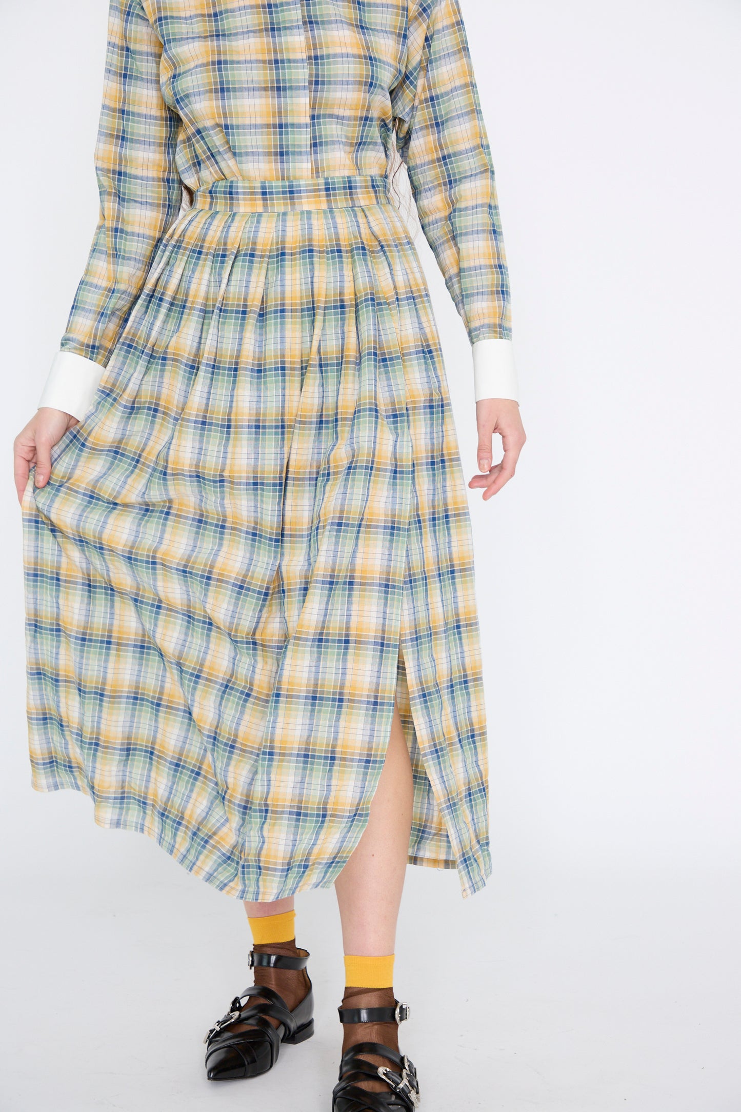 A person dressed in the Jupe Roberta in Paper Miel from Les Prairies De Paris, a plaid cotton dress with long sleeves and a pleated skirt with a slight leg slit, paired with yellow socks and black shoes.