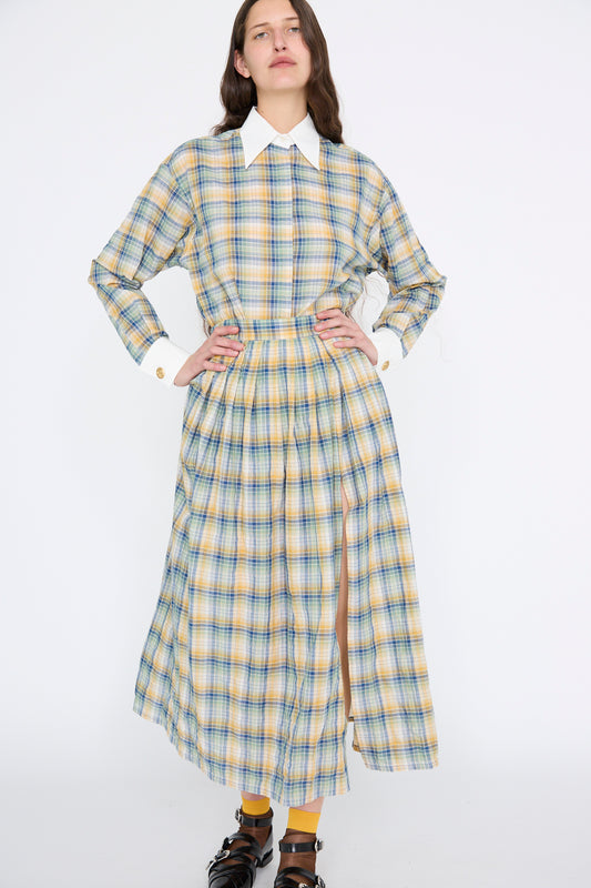 A person is wearing the Jupe Roberta in Paper Miel by Les Prairies De Paris, a long plaid cotton dress with a white collar and cuffs. The sleek front slit enhances its standout style against a plain background.