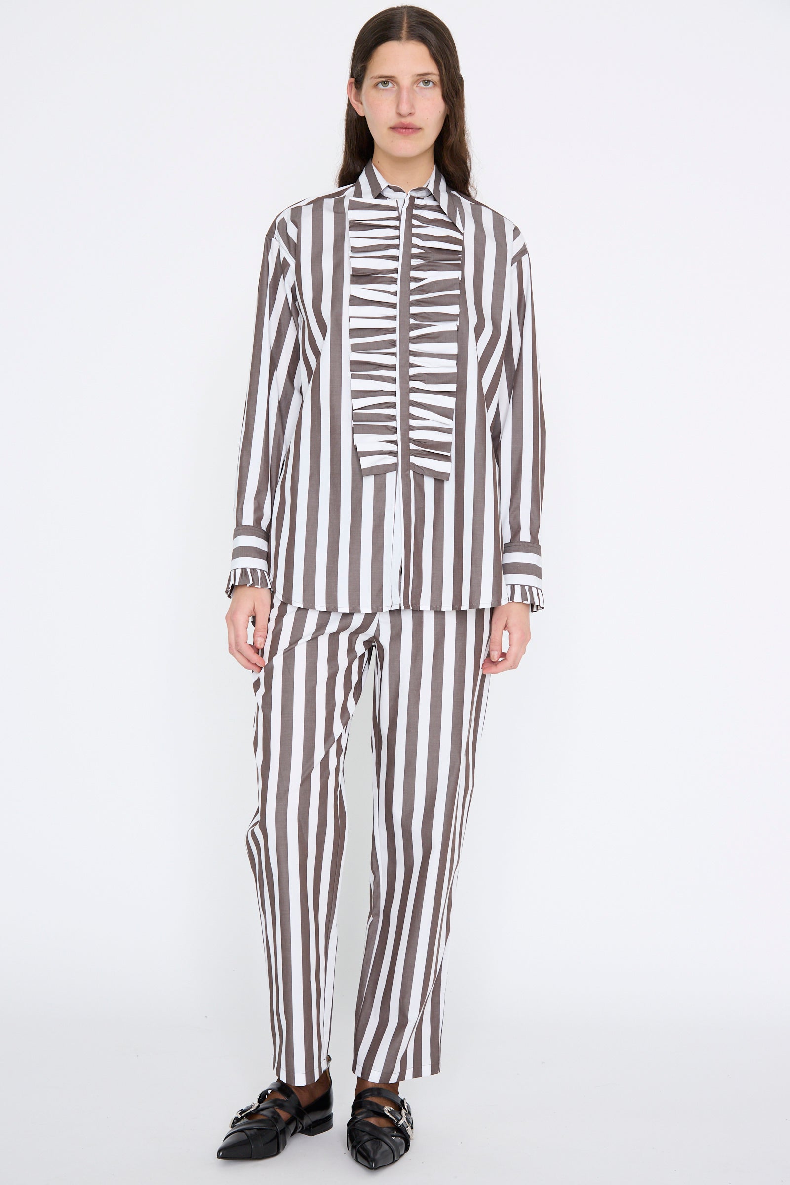 A person wears Les Prairies De Paris’ Pantalon Nicolo in Marron, paired with a black and white striped shirt and sleek black shoes, standing against a plain background.