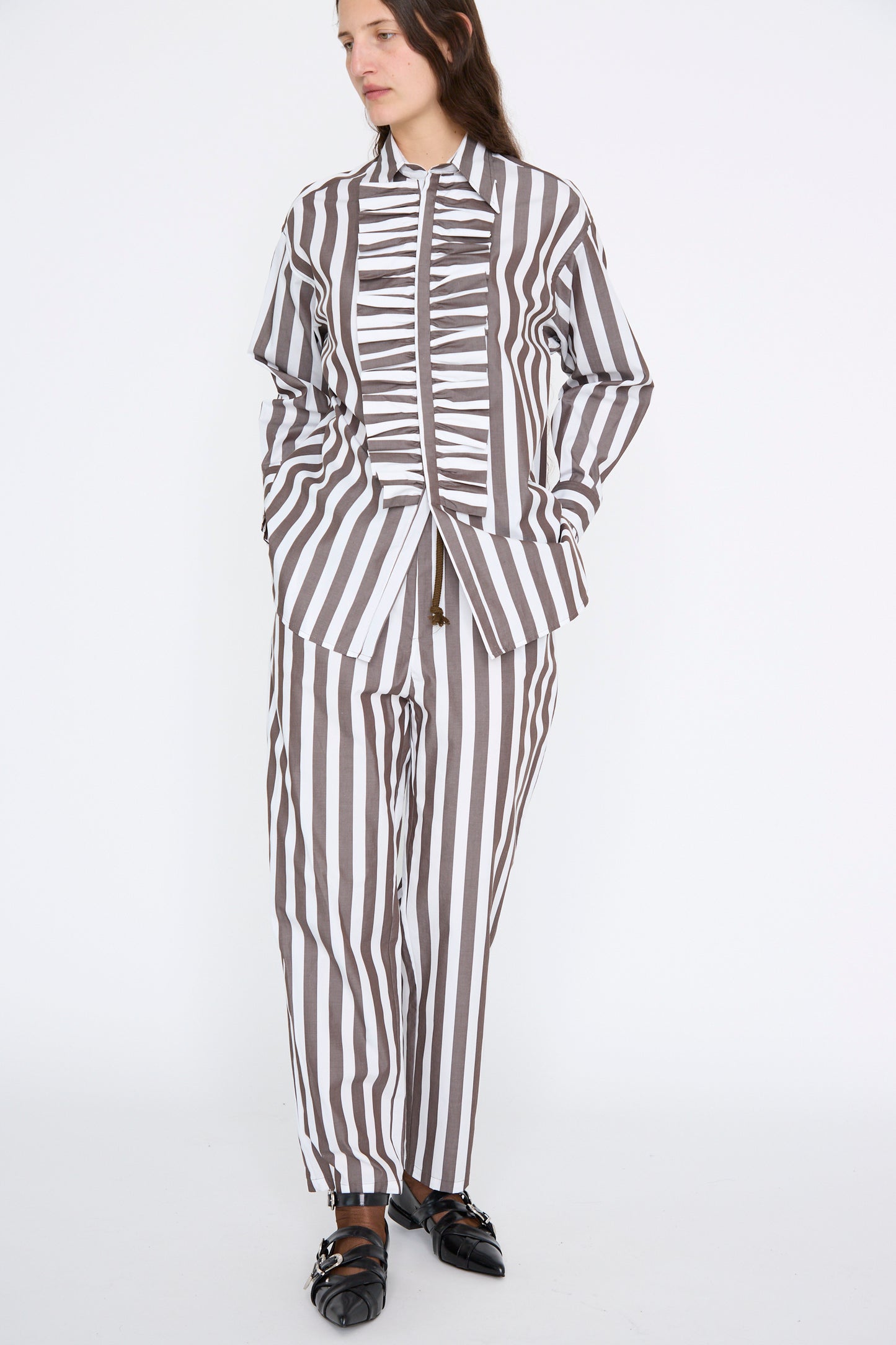 A person stands against a white background in Les Prairies De Paris' Pantalon Nicolo, featuring a relaxed fit, brown and white vertical stripes in organic cotton. The outfit comprises a long-sleeve shirt and matching pants, completed with sleek black shoes.