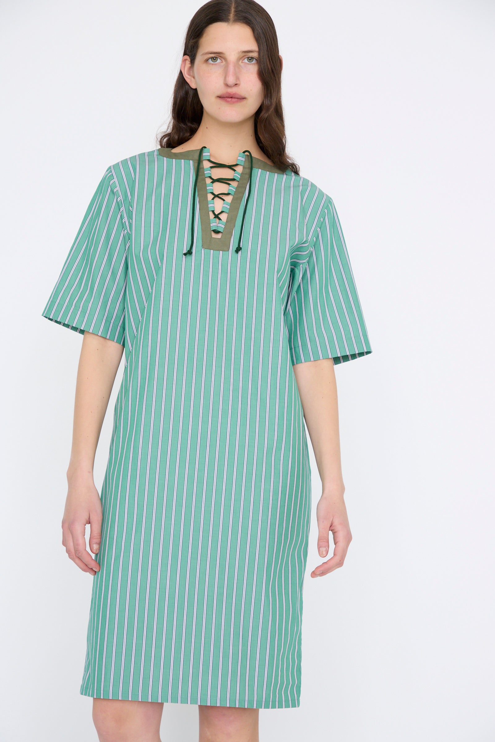 A person wearing the Les Prairies De Paris Robe Giulia in Menthe, a knee-length dress in green and white stripes, features a charming lace-up string tie at the neckline.