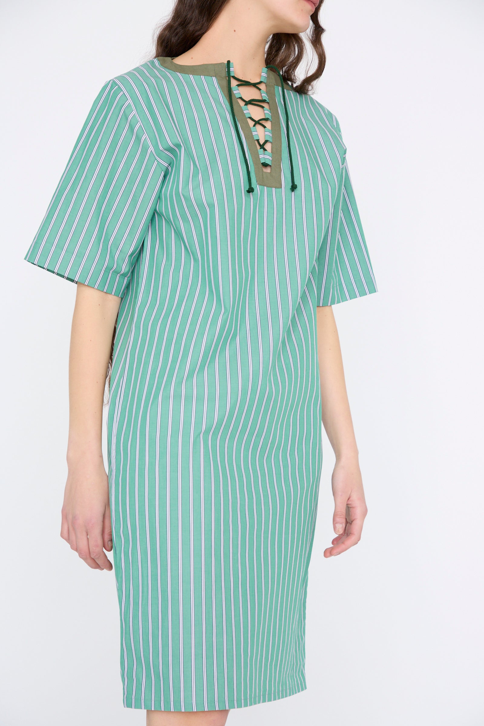 A person is wearing the Robe Giulia in Menthe by Les Prairies De Paris, a green knee-length cotton dress with vertical stripes and a lace-up string tie neckline.