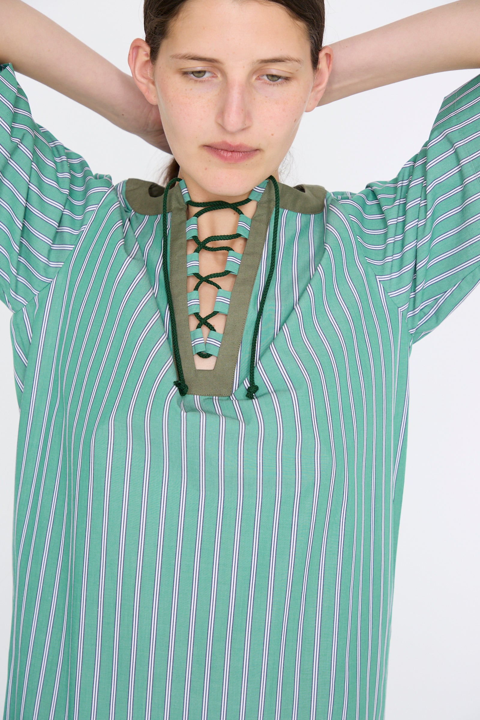 A person wearing the Robe Giulia in Menthe by Les Prairies De Paris, featuring a green and blue striped pattern and a lace-up string tie neckline, gazes downward with hands behind their head against a plain background.