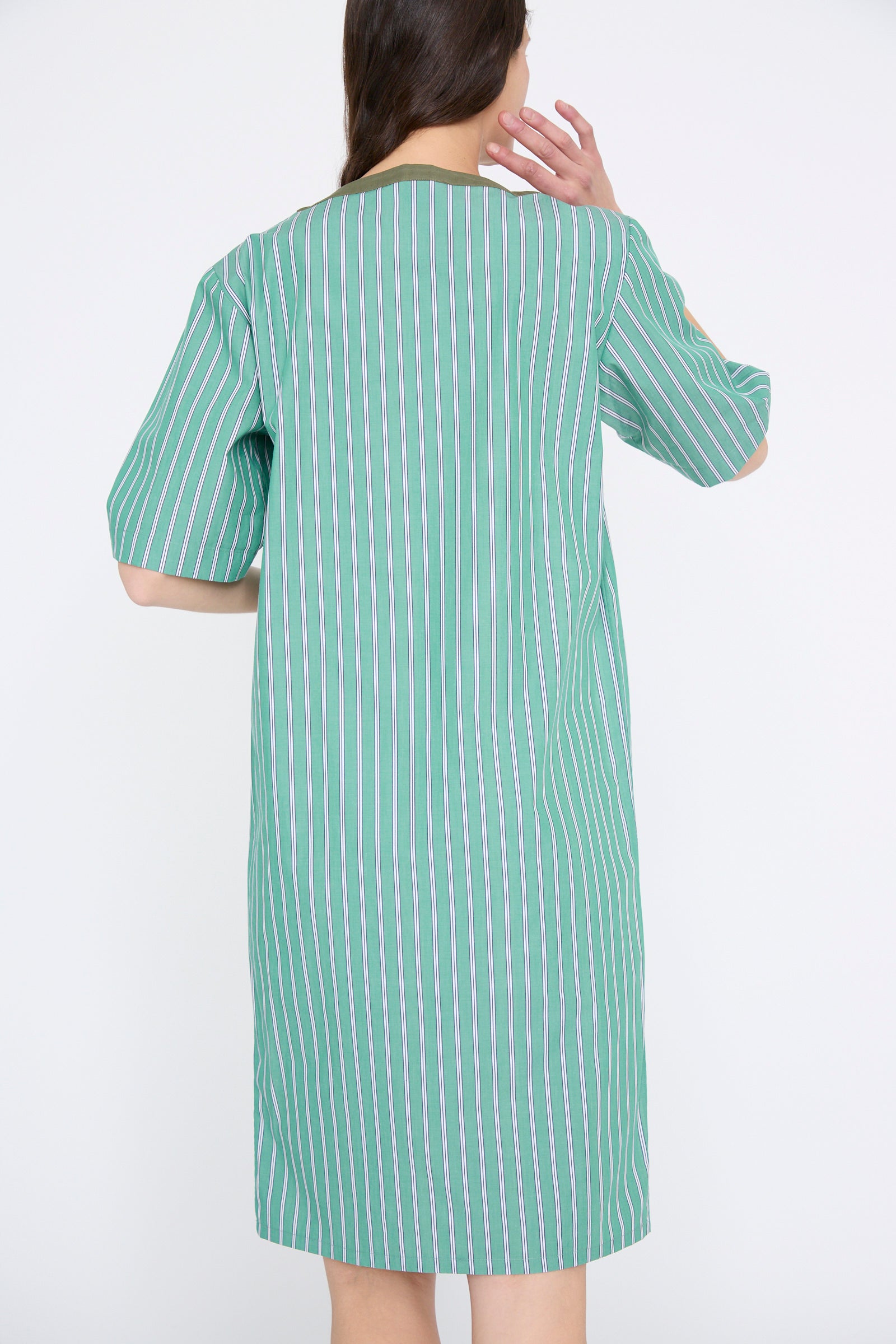 A person with long hair stands facing away, wearing the Robe Giulia in Menthe by Les Prairies De Paris, a green striped cotton dress featuring a lace-up string tie.