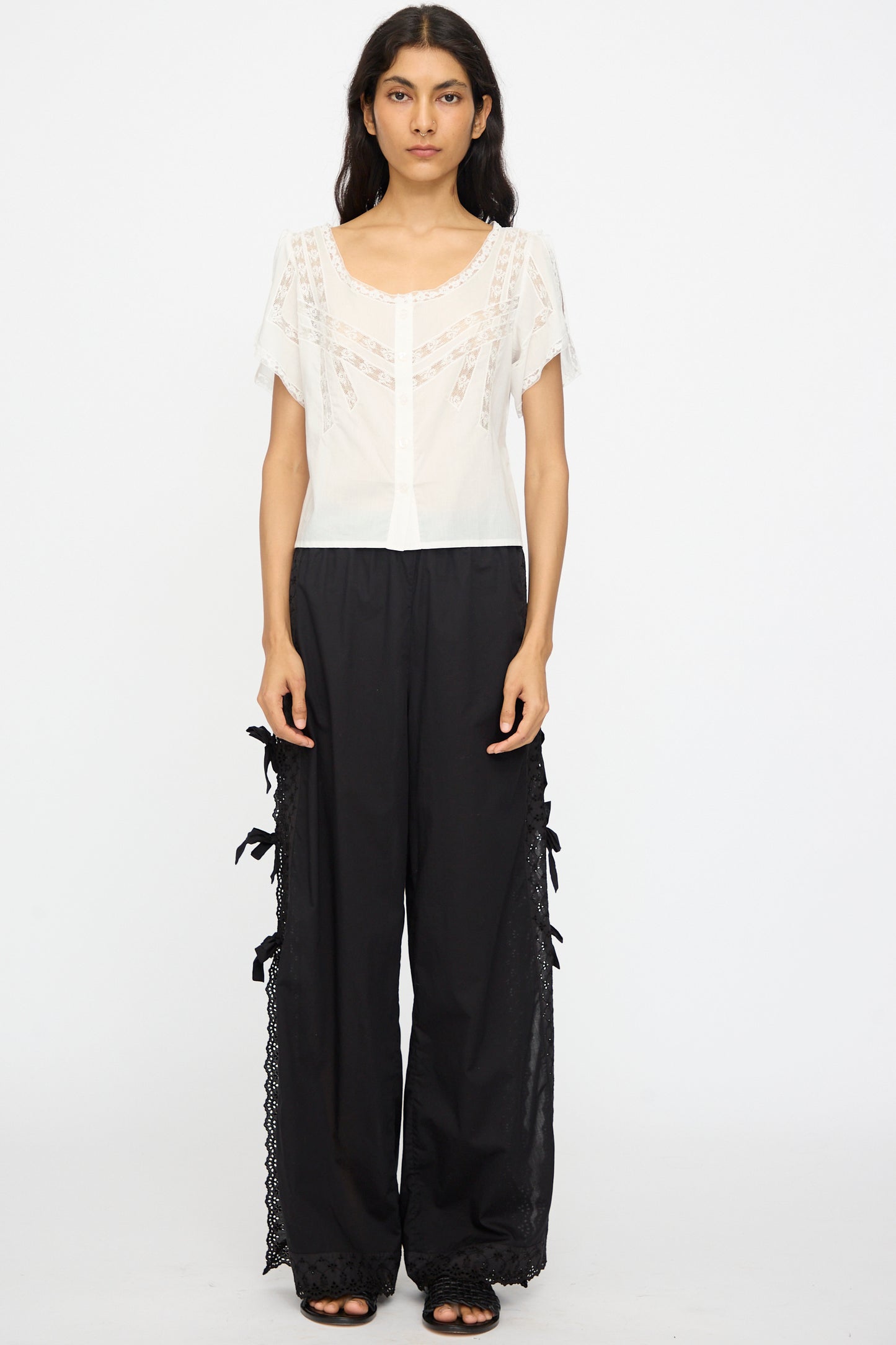A person stands against a plain background in the Loretta Caponi Angela Top in Sunrise, a white lightweight cotton top with lace detailing, styled with black wide-leg pants featuring lace trim and side bows, and black sandals.