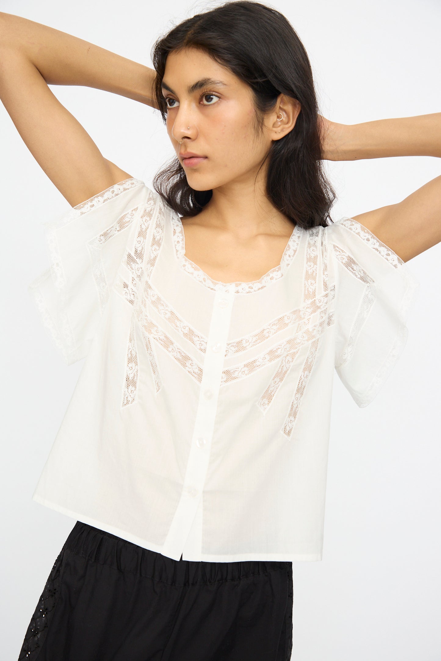 A woman wears the Angela Top in Sunrise by Loretta Caponi—a white lightweight cotton piece with lace details—paired with black pants, standing with her arms raised against a plain background.