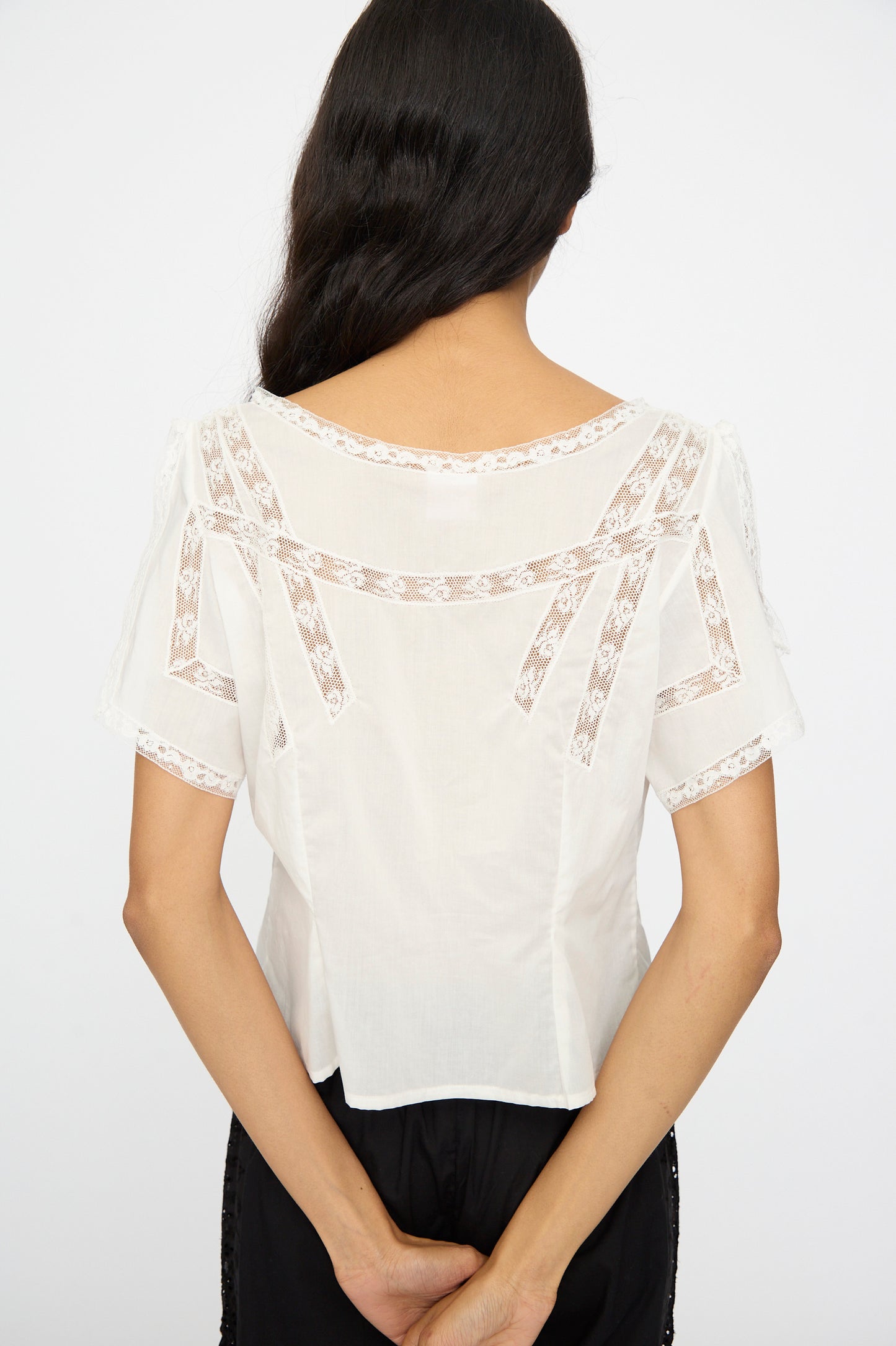 A woman with long dark hair stands facing away, wearing the Angela Top in Sunrise by Loretta Caponi and black pants, her hands clasped behind her back.