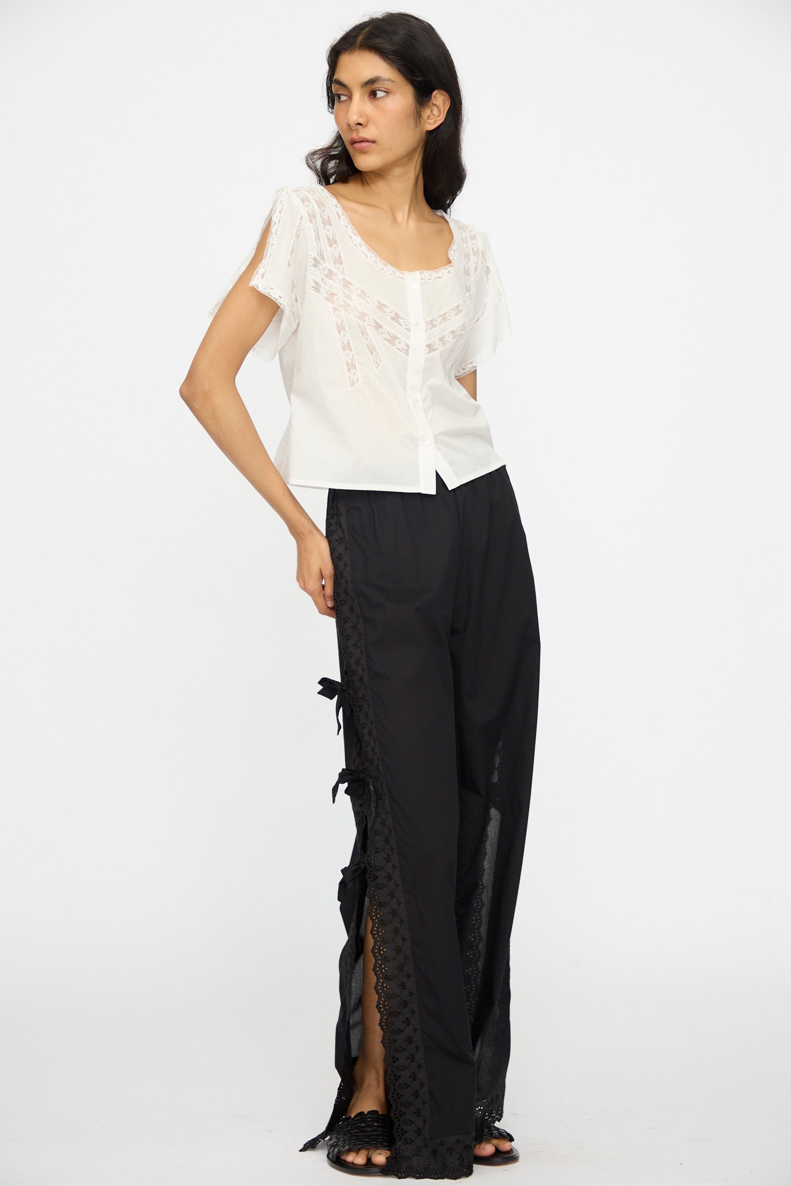 A woman stands against a white background wearing the Angela Top in Sunrise by Loretta Caponi, a lightweight cotton piece paired with loose black pants featuring lace-up and elegant lace details.