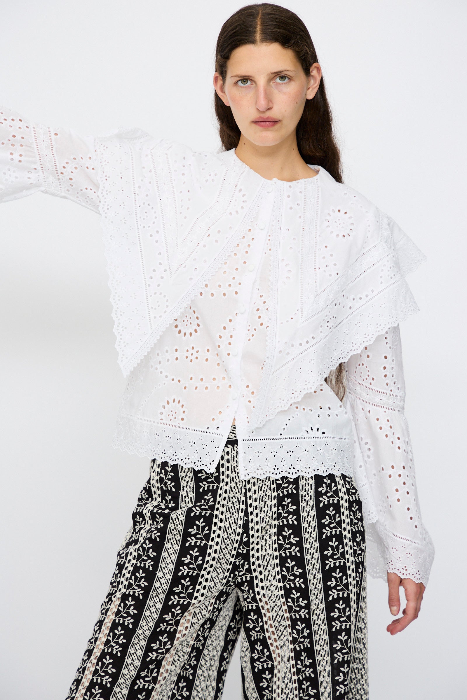 A woman stands against a plain background wearing the Loretta Caponi Azzurra Shirt in Constellation Sangallo, featuring a maxi lace collar and layered details, paired with black-and-white patterned trousers.