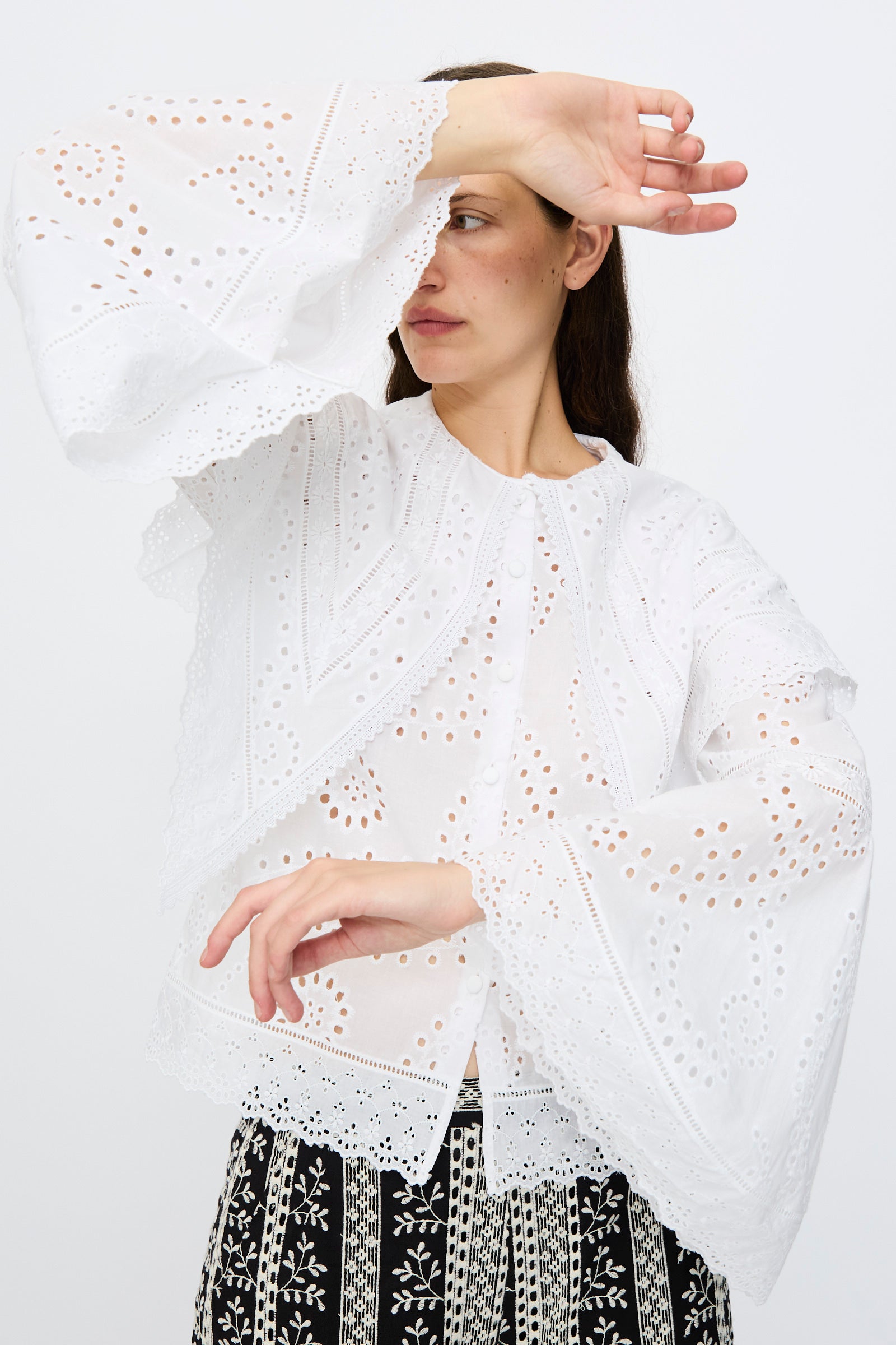 A woman raises one arm, partially covering her face, while wearing the Loretta Caponi Azzurra Shirt in Constellation Sangallo—a white long-sleeved eyelet blouse—paired with patterned black-and-white pants against a plain white background.