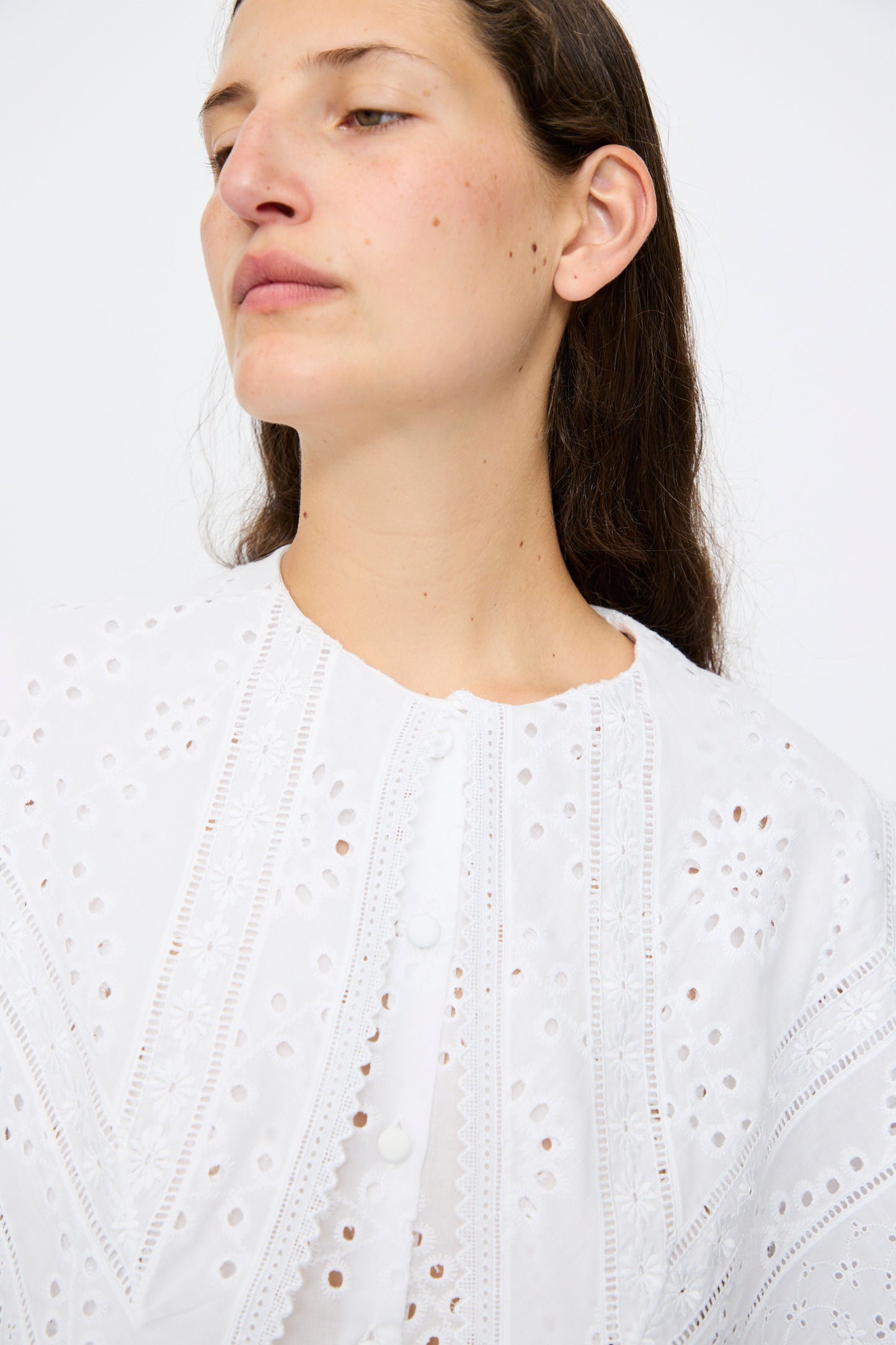 A person with long brown hair wears the Azzurra Shirt in Constellation Sangallo by Loretta Caponi, a white cotton poplin blouse with floral embroidery and delicate eyelet details, looking slightly to the side against a plain background.