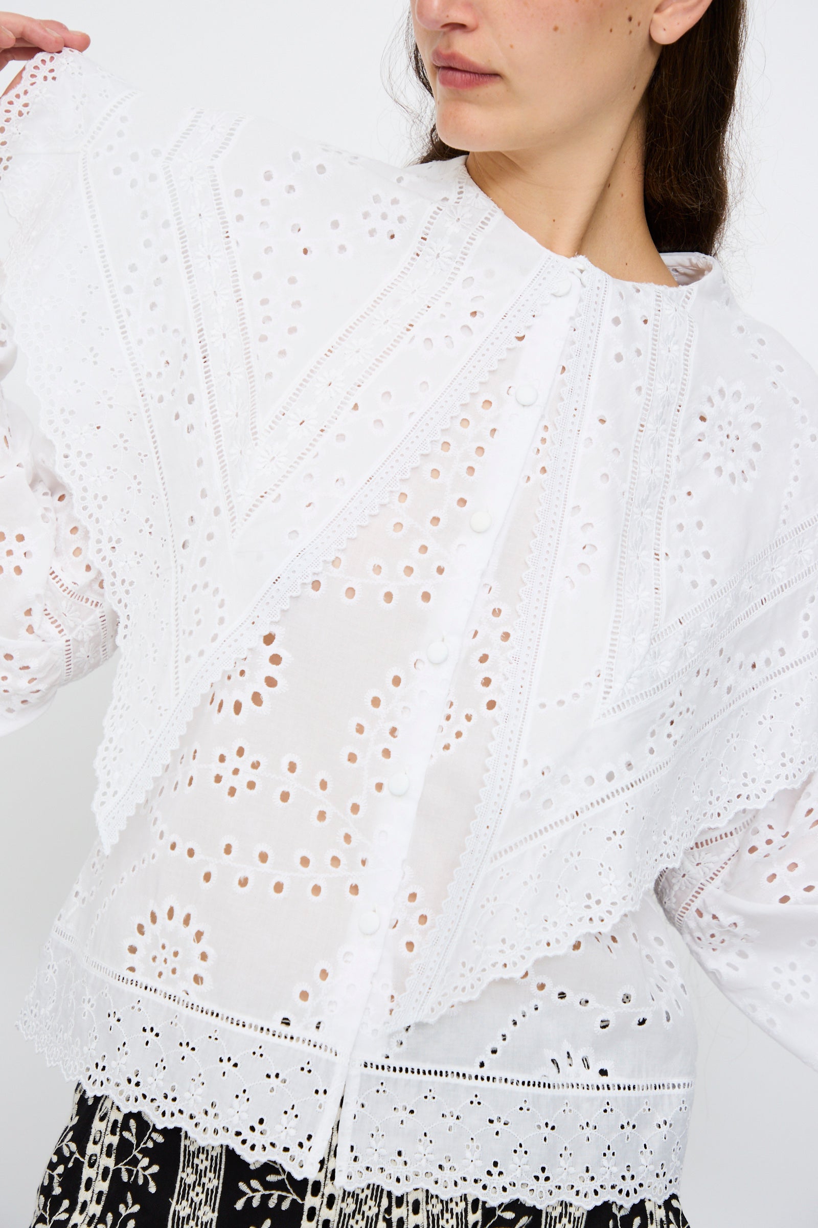 A woman wears the Loretta Caponi Azzurra Shirt in Constellation Sangallo, featuring white long sleeves, eyelet lace with intricate floral embroidery, and scalloped edges. Only her upper body and part of her face are visible.
