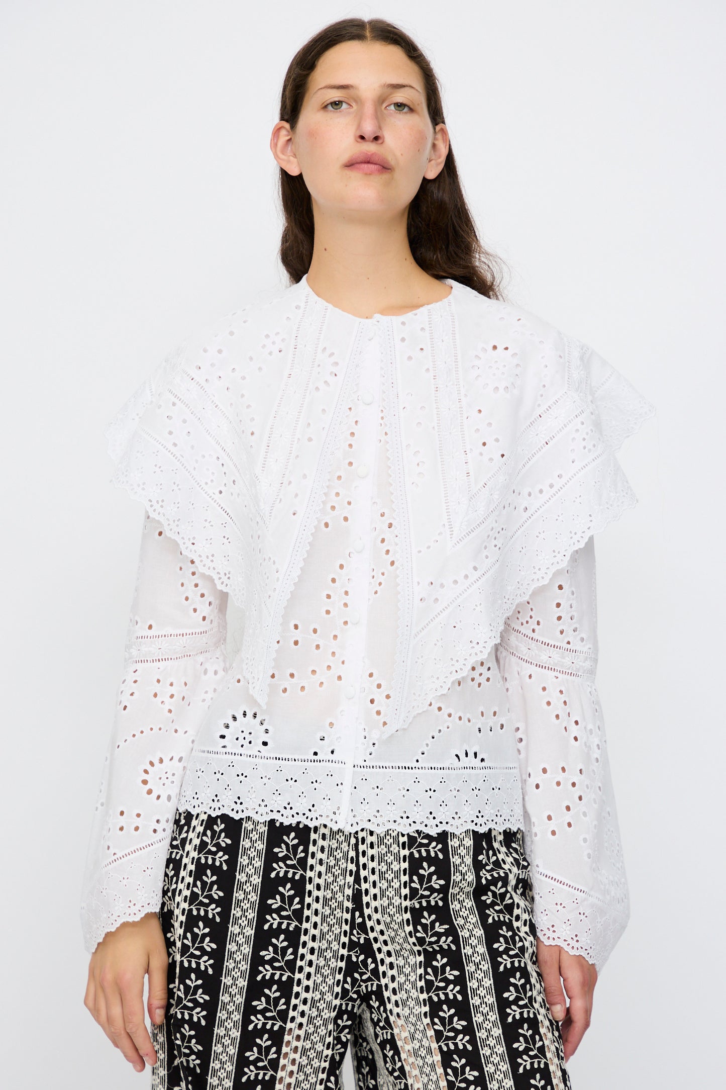 A person wears the Loretta Caponi Azzurra Shirt in Constellation Sangallo, featuring eyelet lace and ruffled details, paired with black patterned pants, standing against a plain white background.