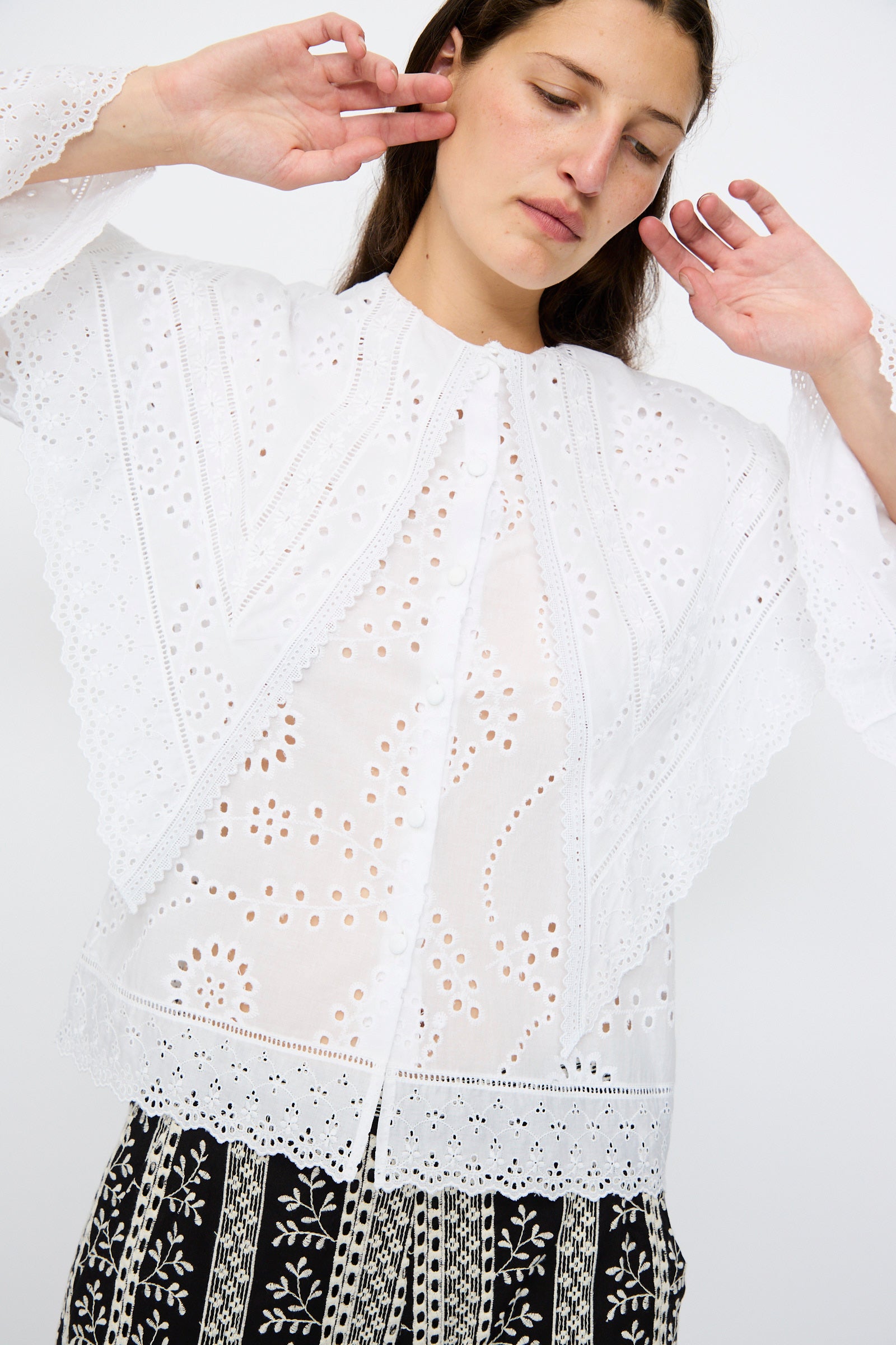 A woman wears the Loretta Caponi Azzurra Shirt in Constellation Sangallo, featuring eyelet embroidery, a maxi lace collar, and scalloped edges, paired with patterned black-and-white pants against a plain backdrop.