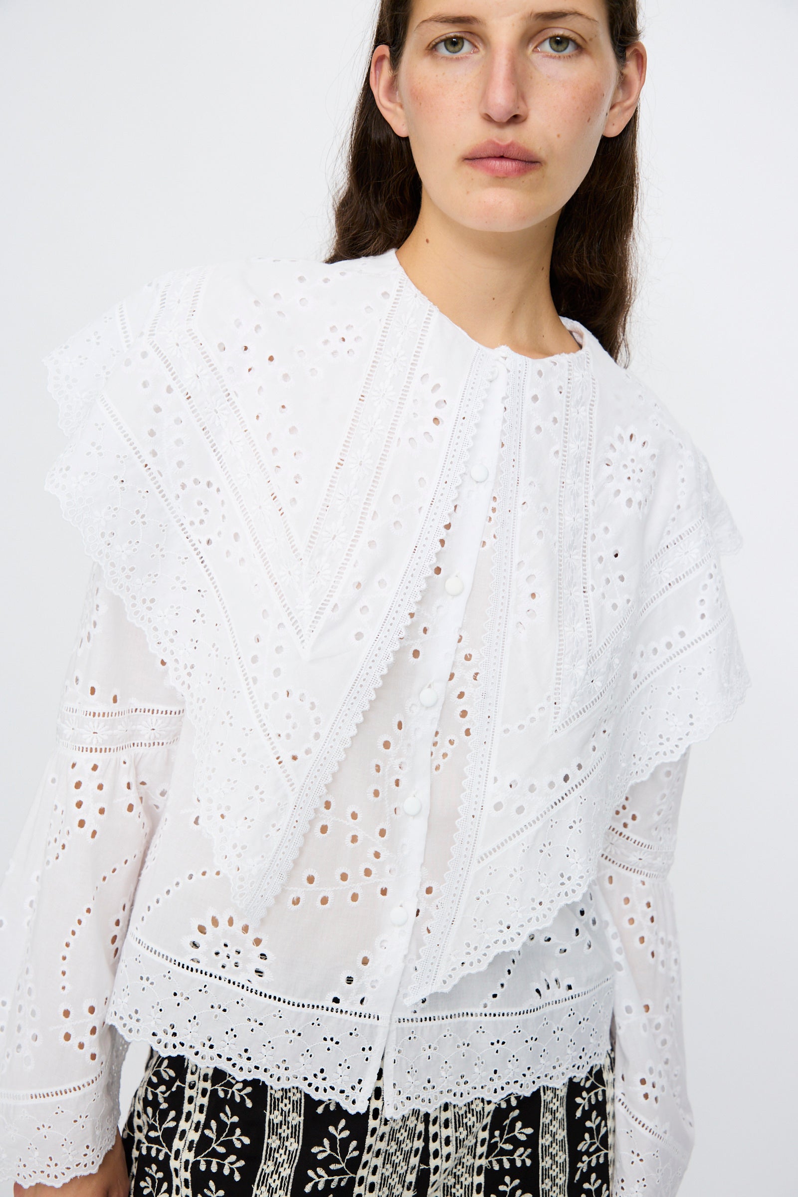 Wearing the Loretta Caponi Azzurra Shirt in Constellation Sangallo, featuring white eyelet lace with a maxi collar and ruffles, paired with patterned black-and-white pants, a person stands against a plain background.