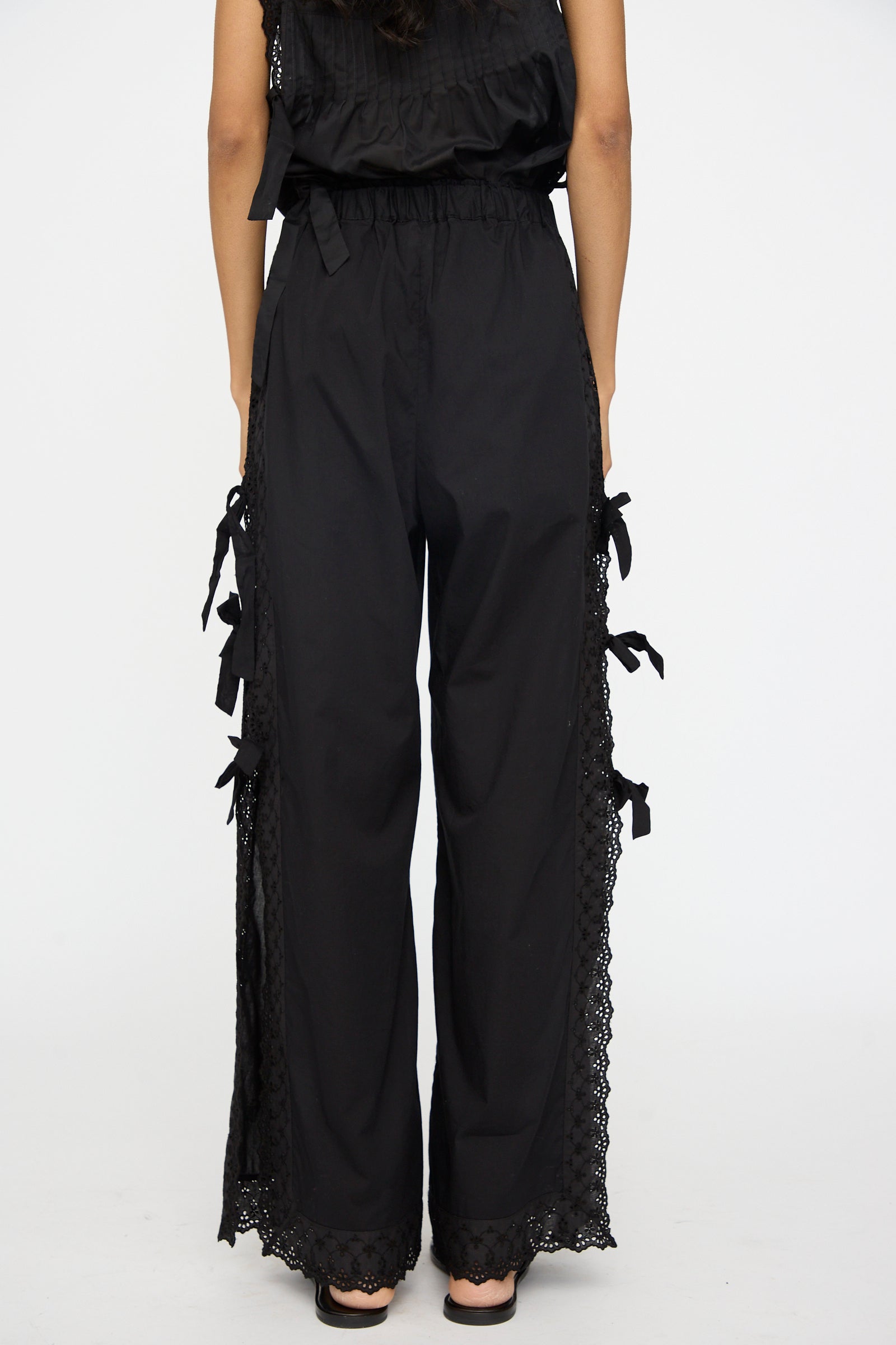 A person models the Clarissa Trousers in Nero by Loretta Caponi, featuring high-waisted black wide-leg pants with lace trim and bow details along the sides, made in Italy, viewed from the back against a plain white background.