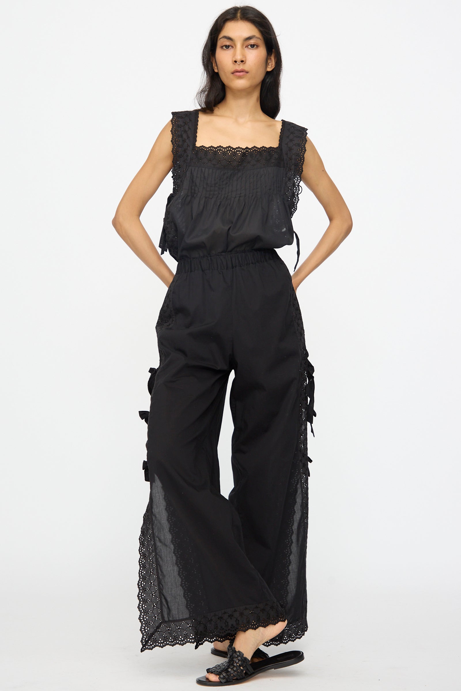 Woman stands against a plain background wearing Loretta Caponi’s Clarissa Trousers in Nero, a high-waisted, trouser-inspired design with lace trim and side bow details, paired with black slide sandals.