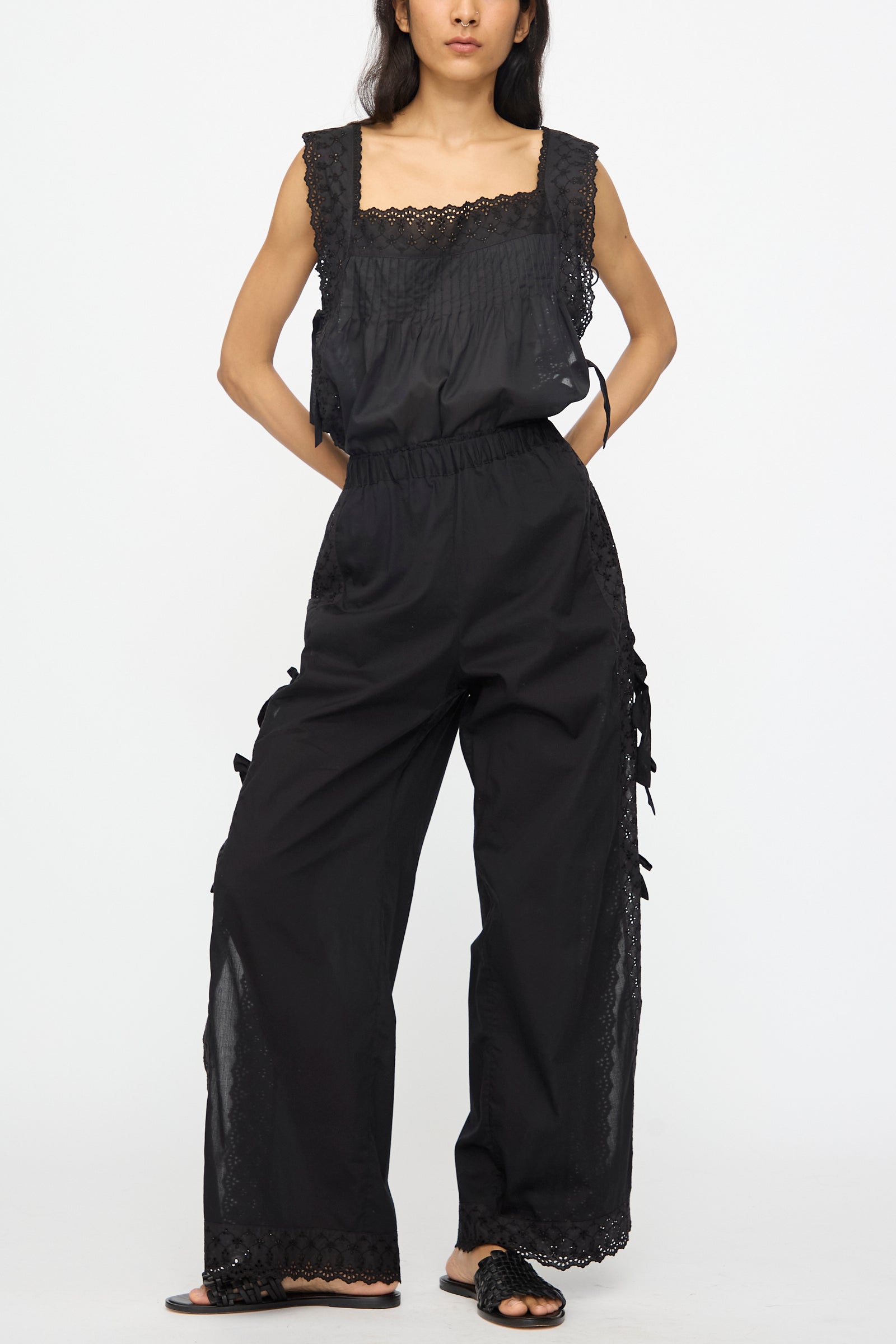 A woman stands wearing Loretta Caponi's Clarissa Trousers in Nero, featuring lace details and bow accents, paired with a black sleeveless top with lace trim and black flat sandals.
