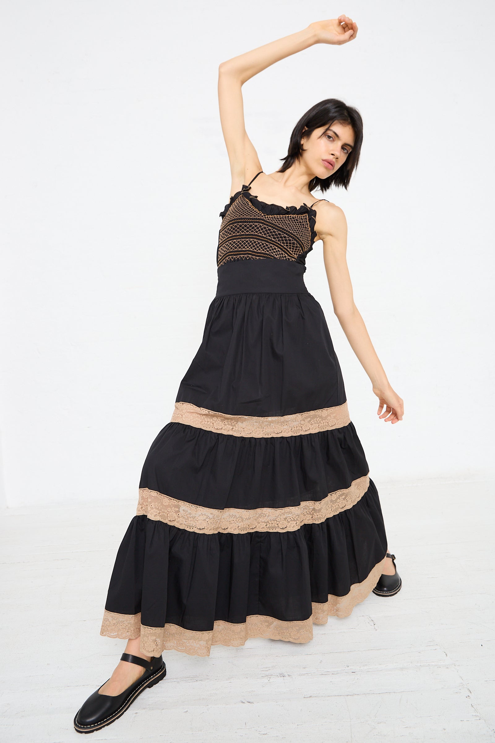 A woman in a black and tan sleeveless dress poses with one arm raised and the other outstretched, standing on a white background. She is wearing black flat shoes and the Loretta Caponi Cotton Violetta Maxi Skirt in Black features a lightweight cotton high waist.