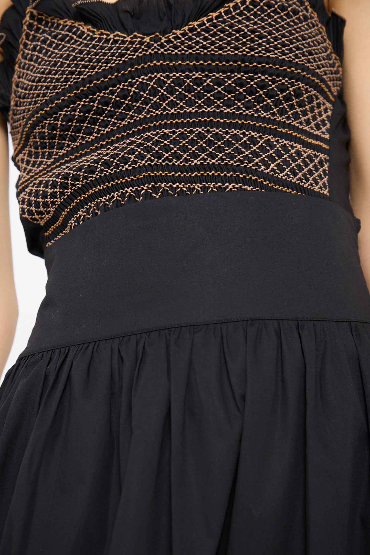 Close-up of a person wearing a black sleeveless dress with a smocked design featuring intricate orange stitching on the upper portion and a solid black Cotton Violetta Maxi Skirt in Black by Loretta Caponi.