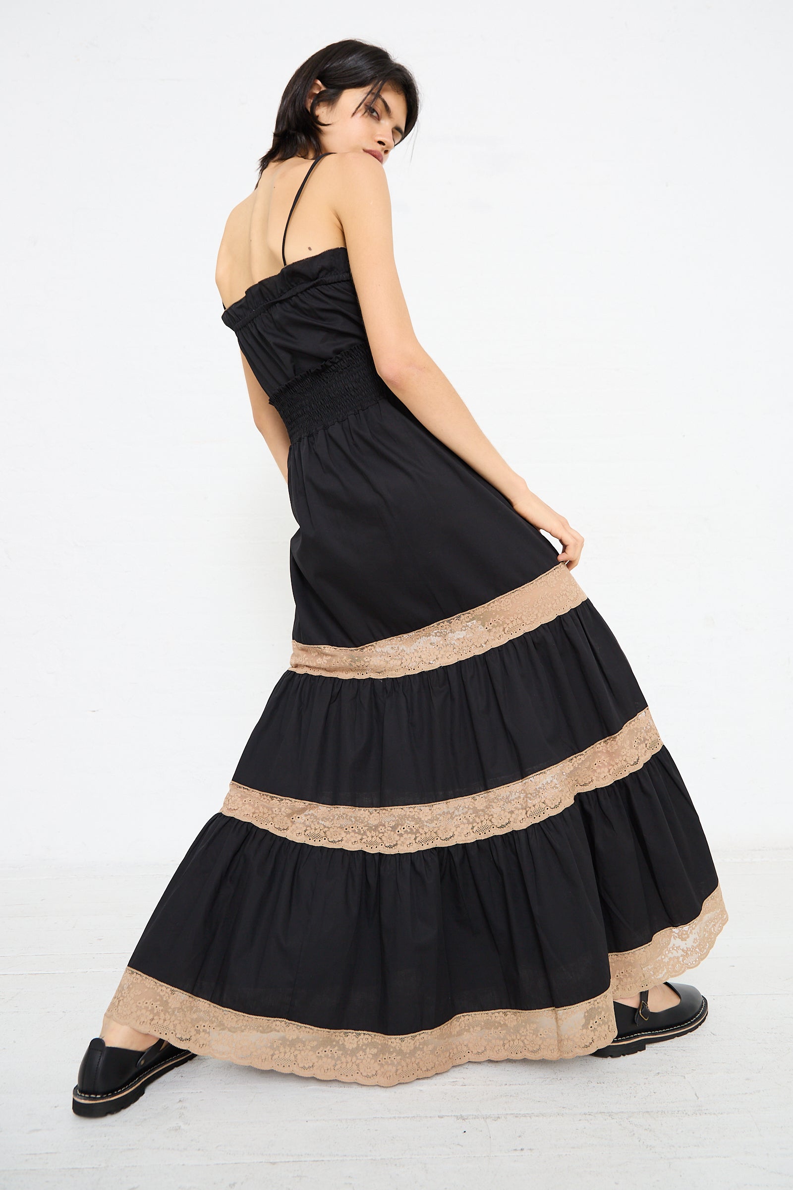 Woman in a black Cotton Violetta Maxi Skirt by Loretta Caponi with beige lace trim at the hem, standing against a white background. She is turned slightly to her left and looking over her shoulder, the high waist design accentuating her elegant silhouette.