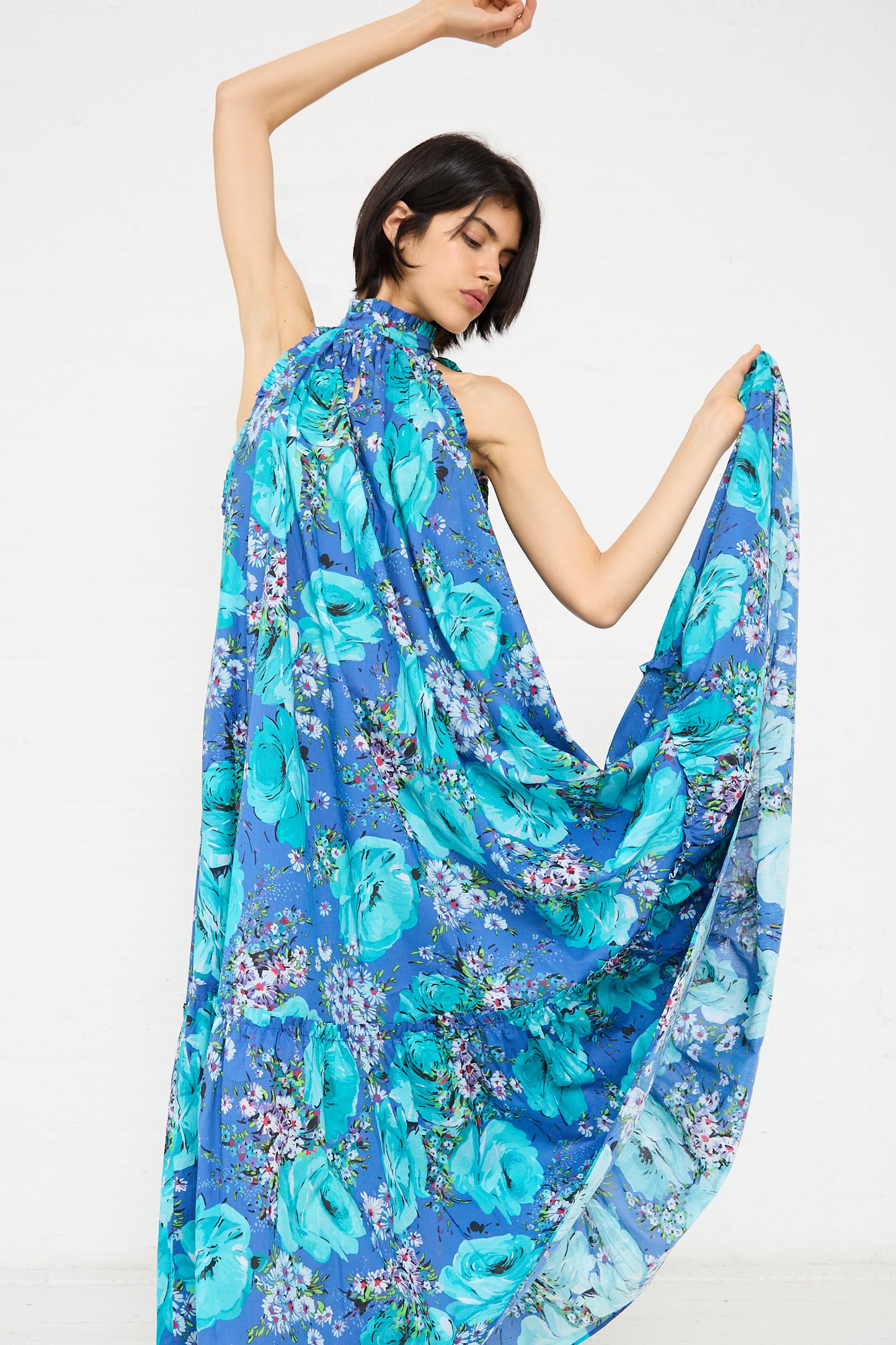 A person in a sleeveless Loretta Caponi Melinda Dress in Sky of Roses adorned with a floral pattern poses with one arm raised overhead and the other arm holding out one side of the garment, creating a flowing, draped effect.