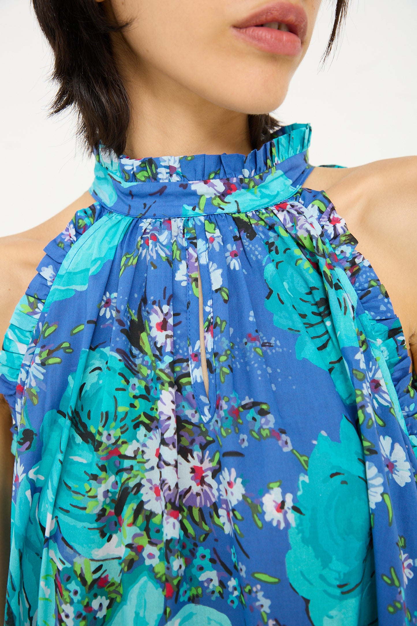 A close-up of a person wearing the Loretta Caponi Melinda Dress in Sky of Roses with a ruffled neckline and multi-colored floral patterns. The person's face is partially visible, and they pair the dress effortlessly with a tiered skirt that complements the vibrant hues.