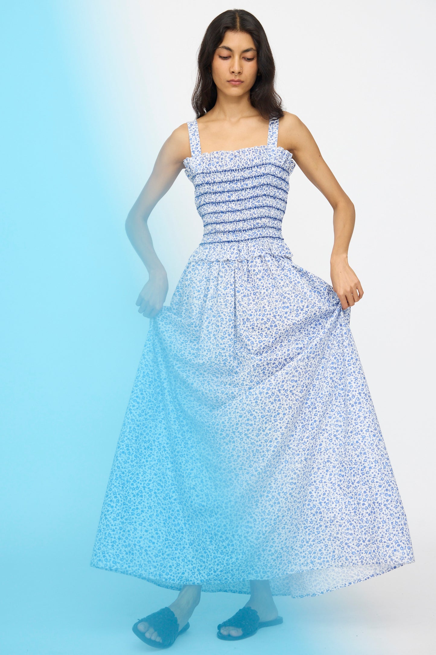 A woman wearing the Loretta Caponi Ombretta Dress in Marina Grande poses against a light background, holding out the sides of her skirt as a blue light partially covers the lower left side of the image.