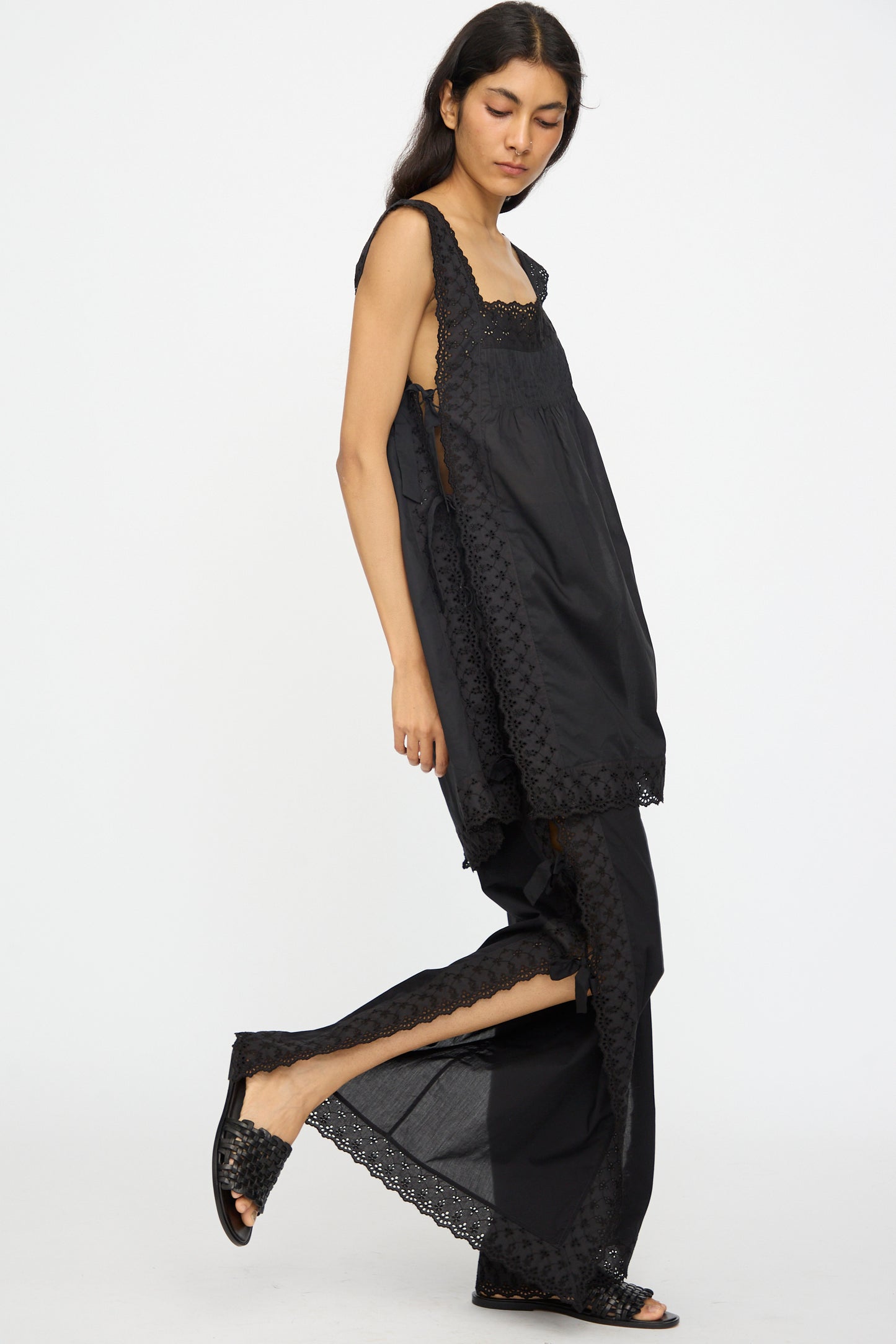 A person stands sideways against a plain white background wearing the Loretta Caponi Oriana Top in Nero, a loose sleeveless black lace-detail tank, paired with matching wide-leg pants and open-toe shoes.