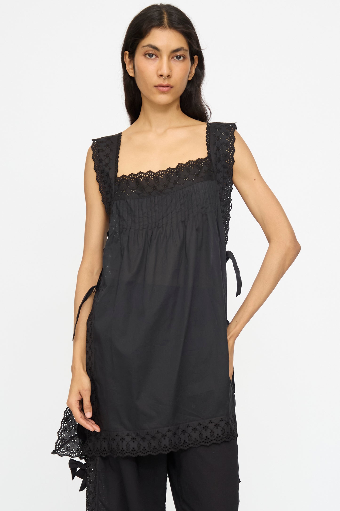 A woman stands against a plain background wearing the Oriana Top in Nero by Loretta Caponi, featuring black lace details and trim, paired with black pants.