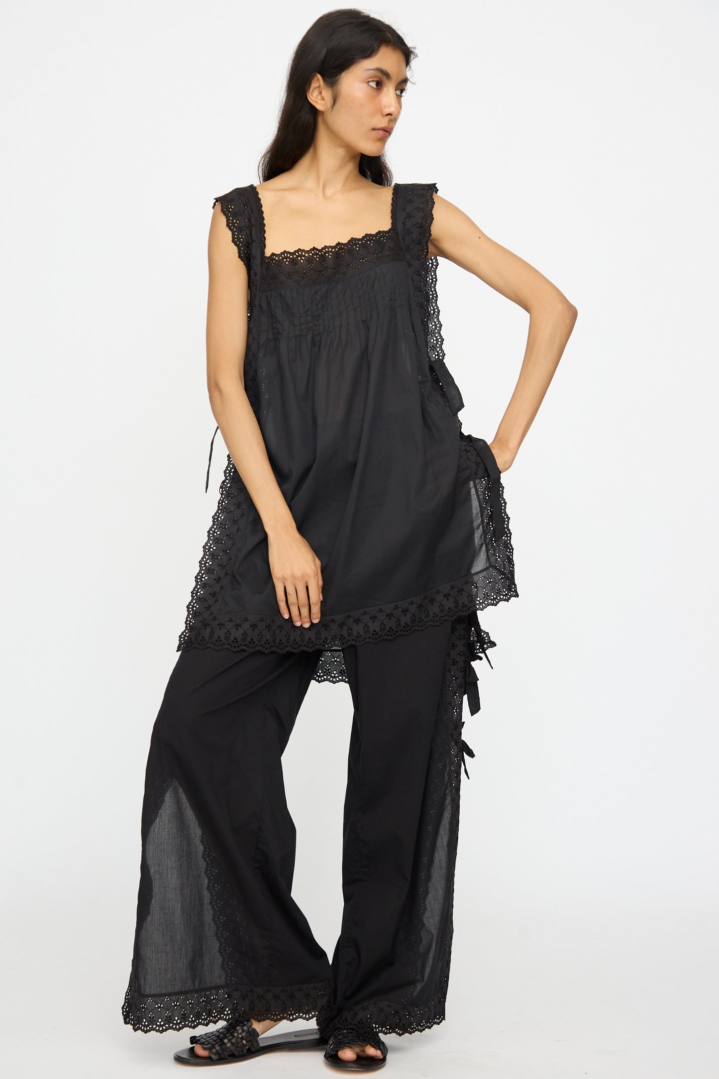 A woman wears the Loretta Caponi Oriana Top in Nero, a sleeveless black lace detail tank, paired with matching loose black pants, looking to the side with one hand on her hip.