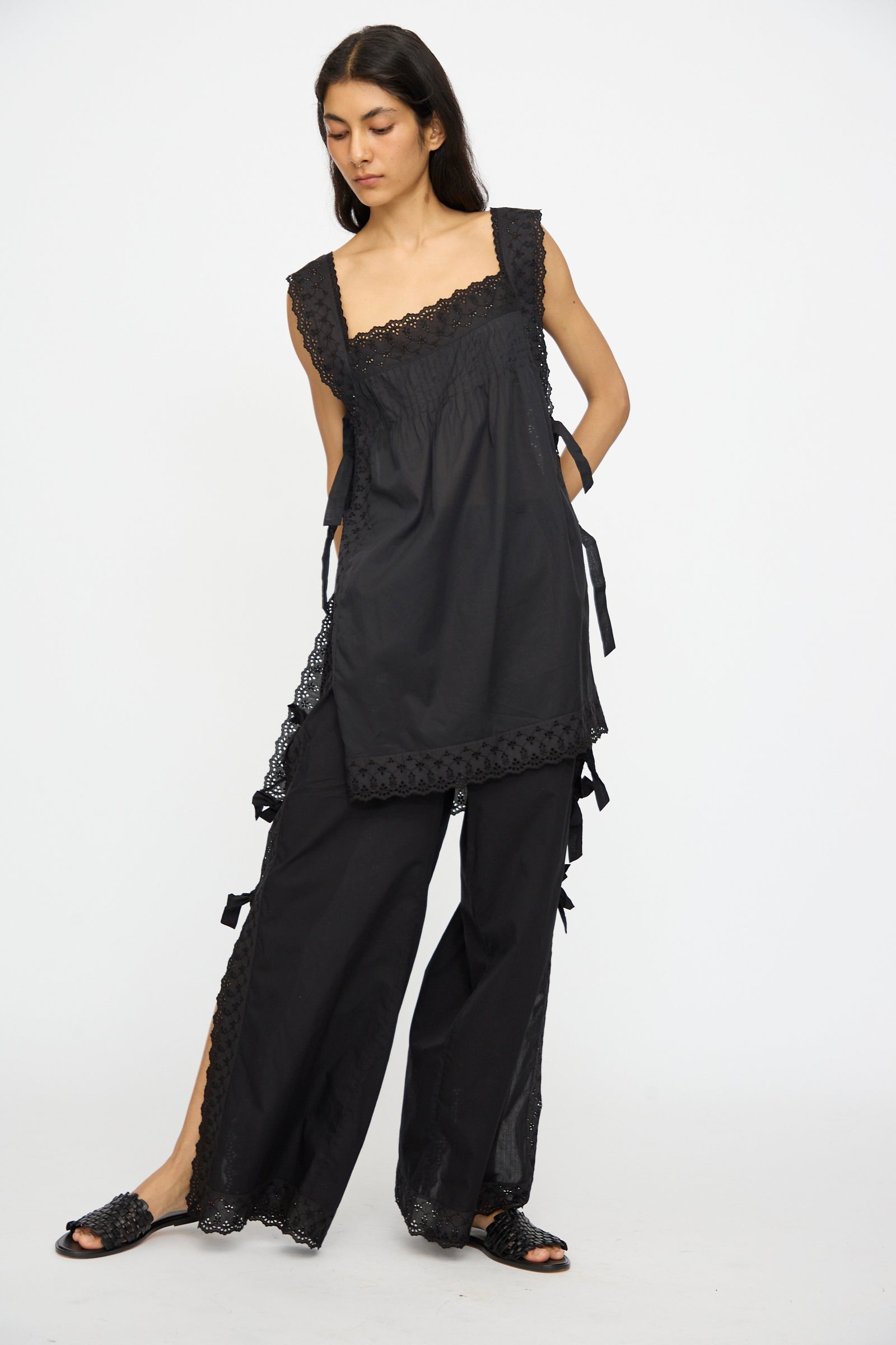 A person poses against a plain white background wearing the Loretta Caponi Oriana Top in Nero, featuring lightweight cotton with lace trim, paired with matching wide-leg pants with lace details and black slip-on shoes.